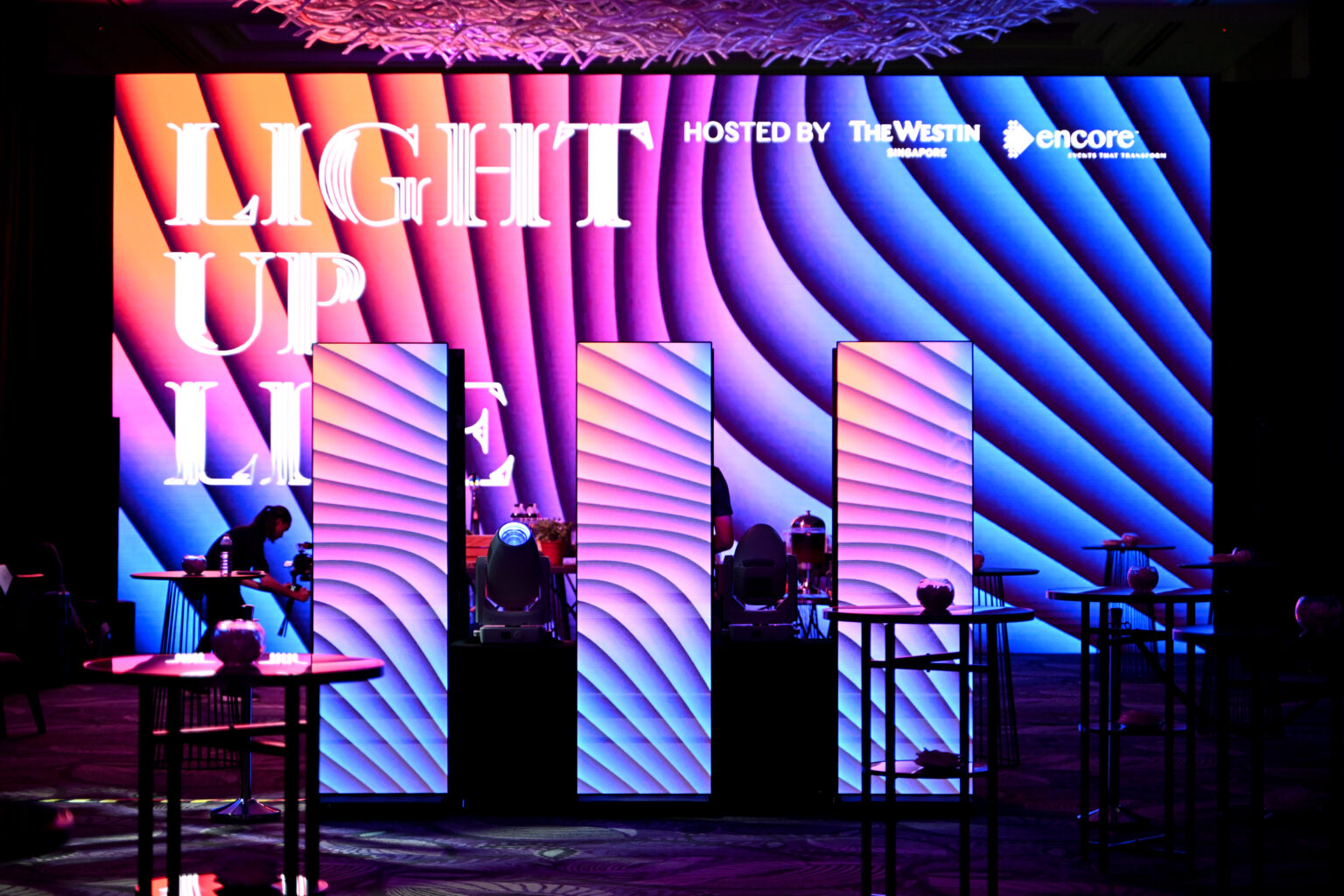 เวทีสีสันสดใสพร้อมรูปแบบแสงสีฟ้าและสีม่วงหยัก และเสาไฟส่องสว่างสามต้นยืนอยู่หน้าป้ายขนาดใหญ่ที่เขียนว่า 