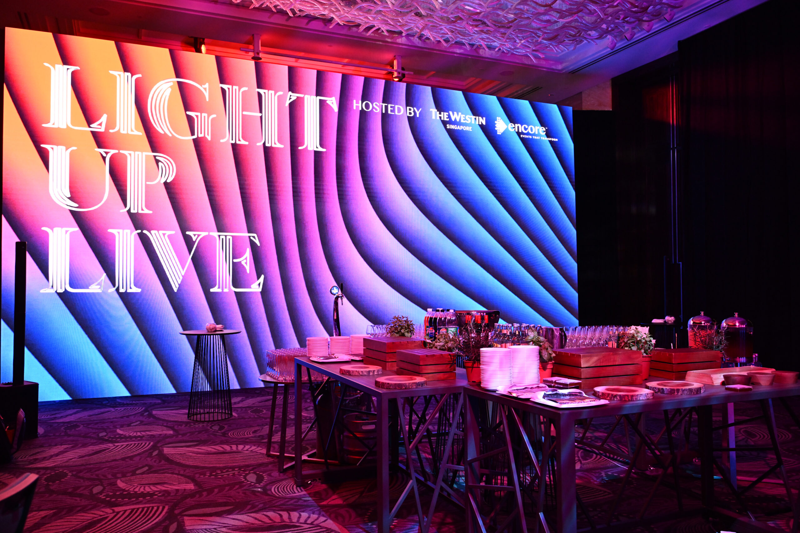Light Up Live: A Stellar Showcase at The Westin Singapore... A colorful event space features a large LED screen displaying “LIGHT UP LIVE” beside a table set with food, drinks, plates, and utensils under dramatic pink and blue lighting.