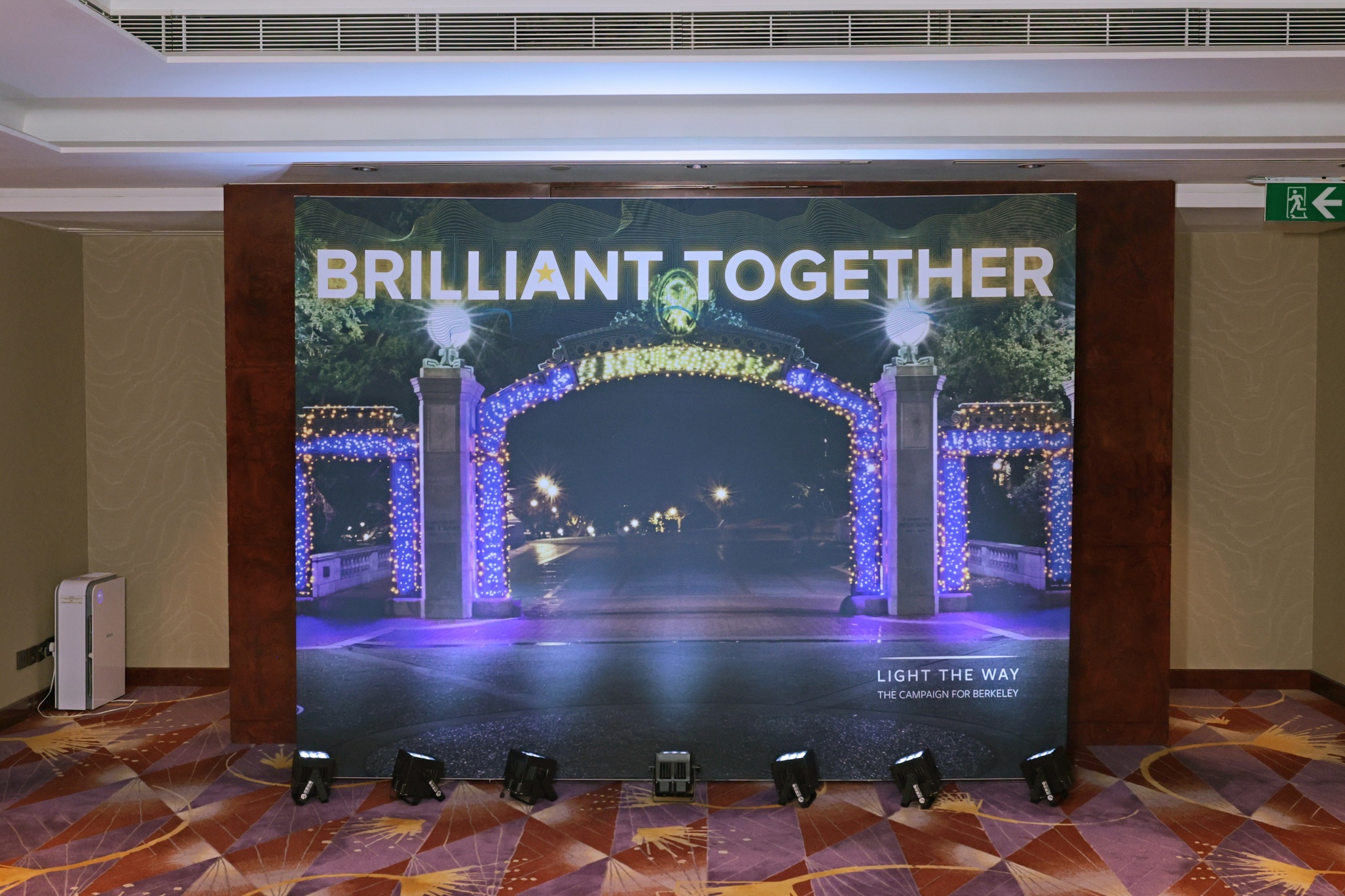 A large indoor display features a nighttime photo of a lit archway with the text BRILLIANT TOGETHER at the top and LIGHT THE WAY THE CAMPAIGN FOR BERKELEY at the bottom right. Several spotlights are placed in front.