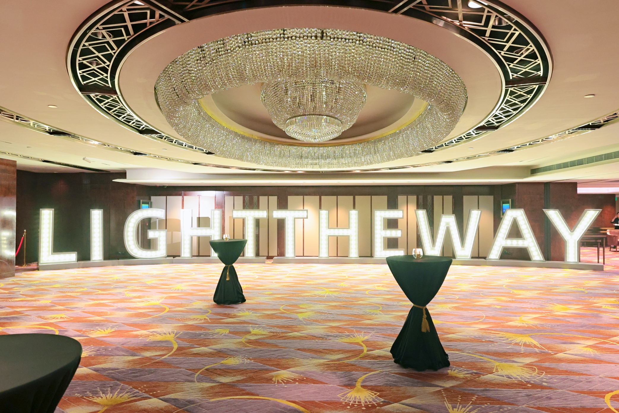 A spacious, elegant ballroom features a large chandelier and illuminated letters spelling LIGHT THE WAY. Three tall cocktail tables with dark covers are arranged on a patterned carpet.