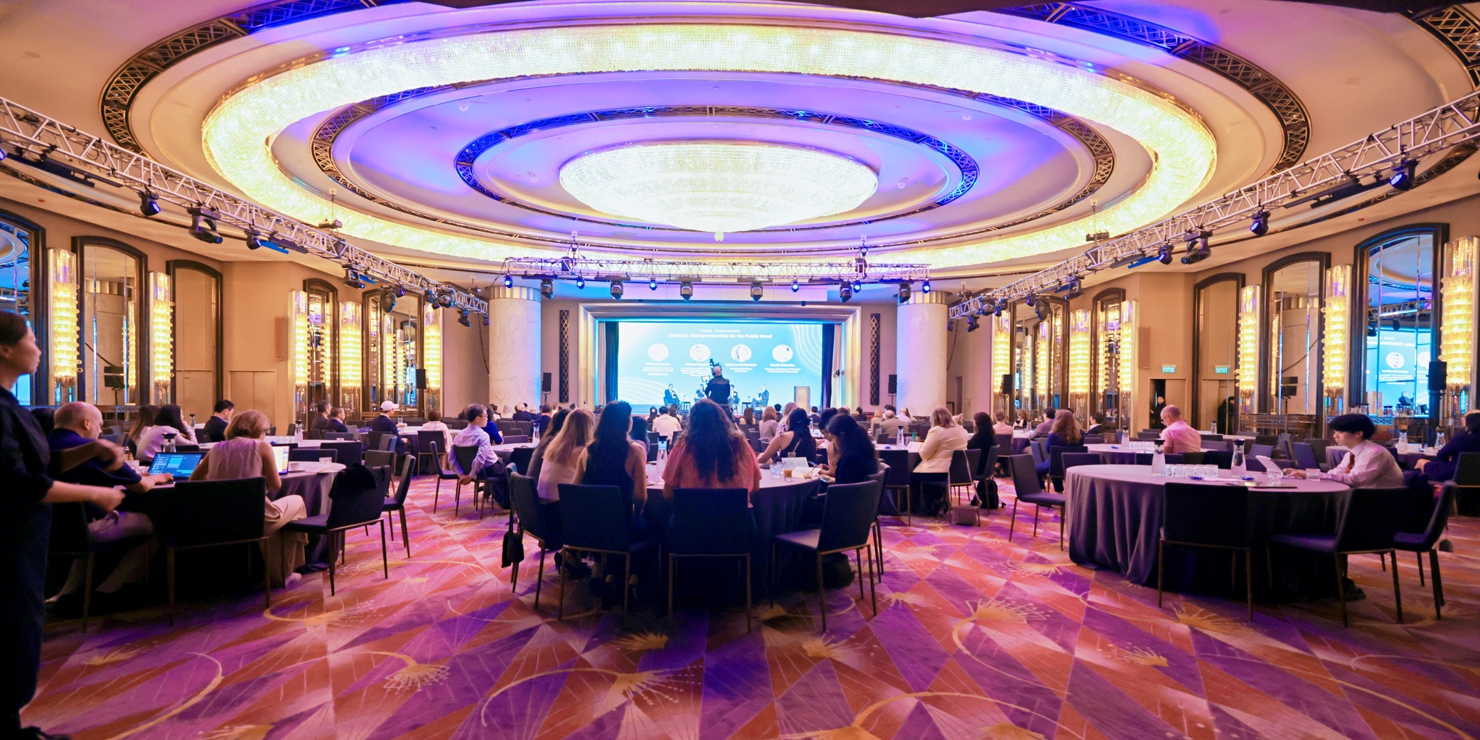 A large conference room with round tables and attendees seated, facing a brightly lit stage with a projection screen under an ornate ceiling with circular lights.