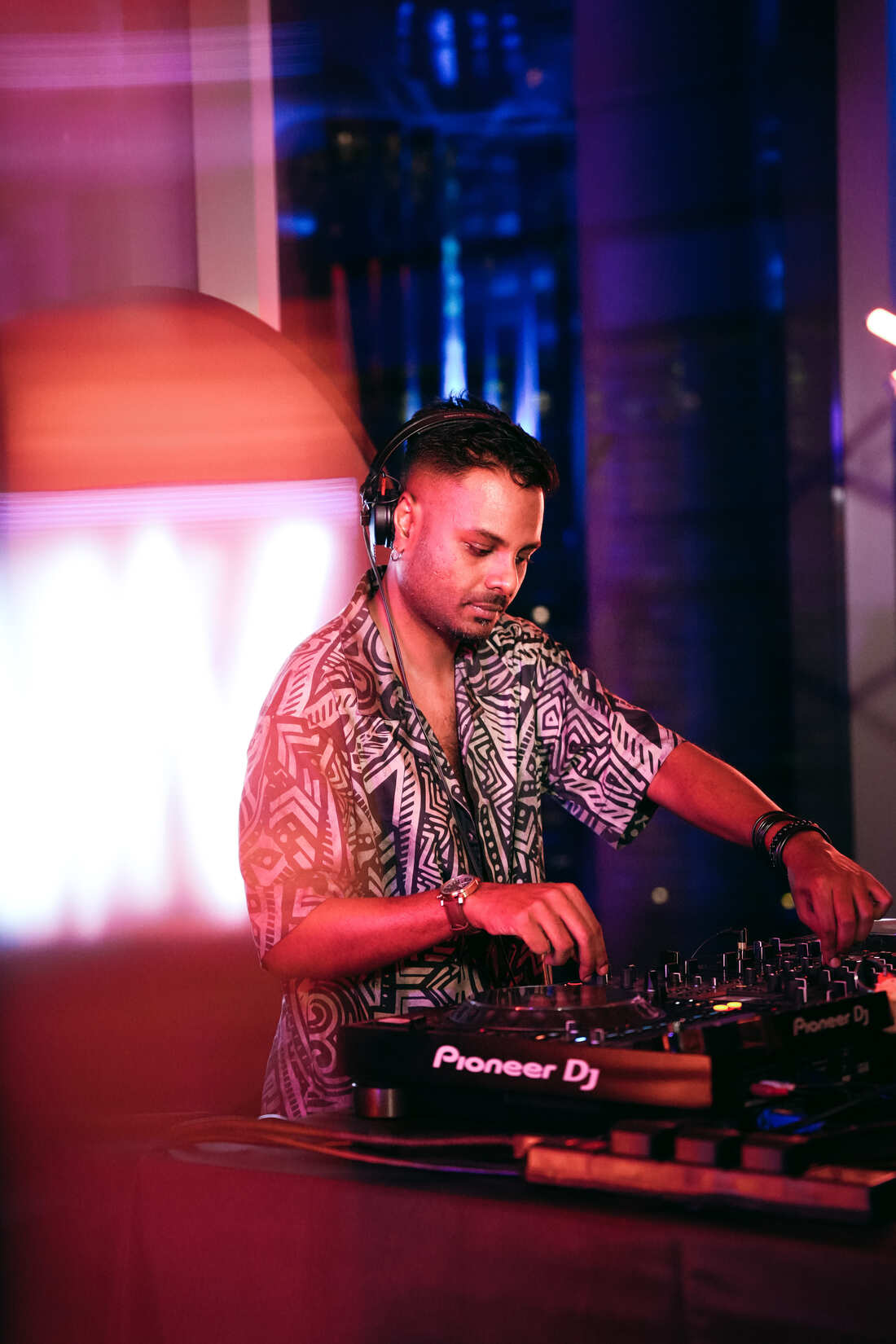 A DJ wearing headphones and a patterned shirt mixes music on a Pioneer DJ controller at a colorful, dimly lit event with vibrant pink and blue lighting effects.