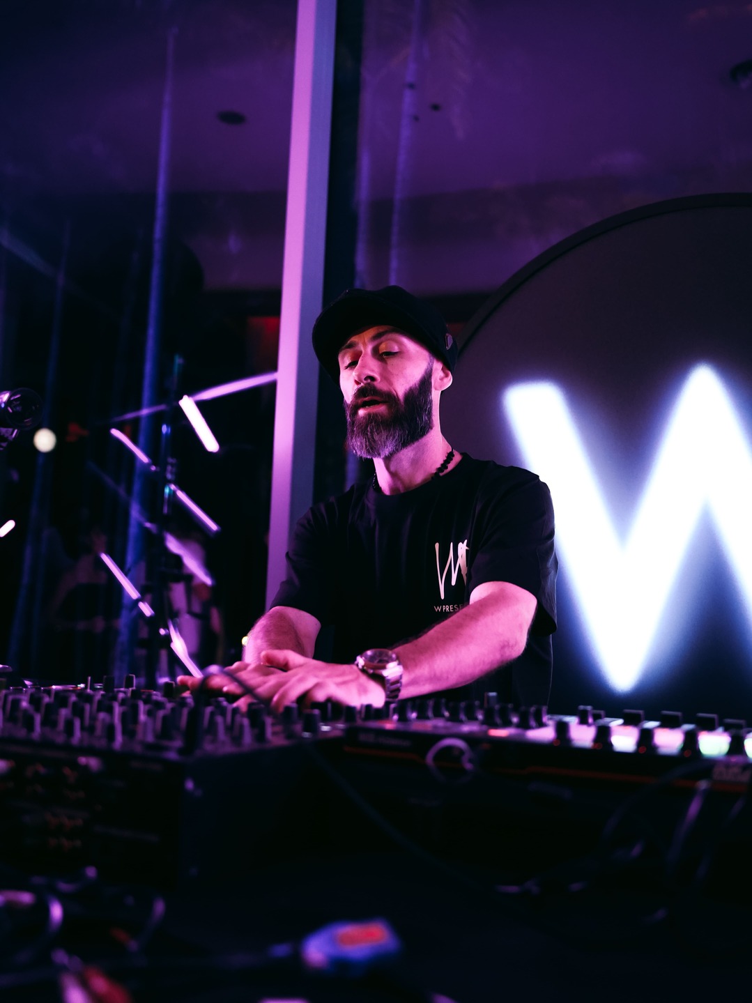 A bearded DJ wearing a black t-shirt and cap performs behind a mixer, illuminated by purple lighting, with a large glowing W in the background.