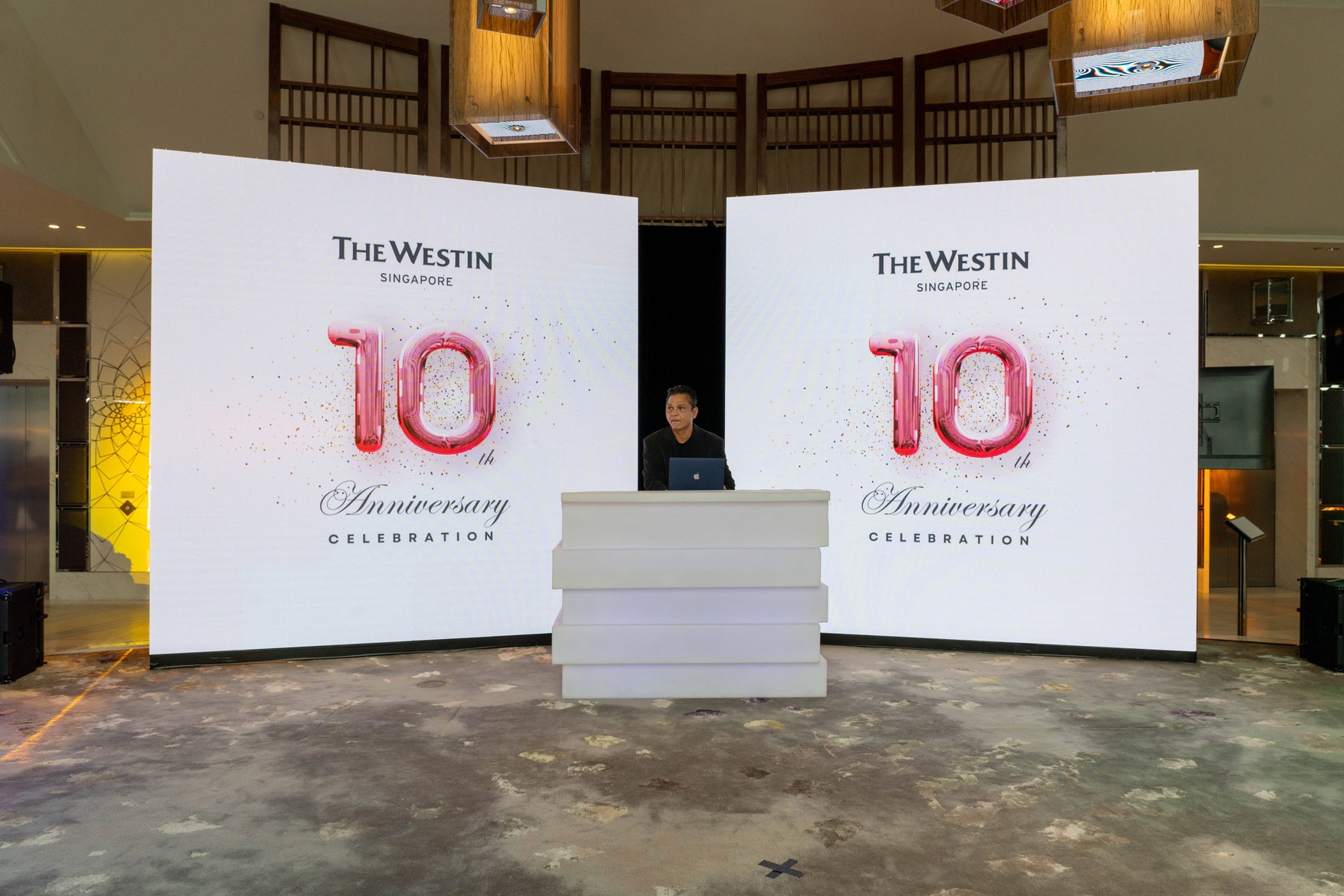 A person stands behind a white podium between two large digital screens displaying The Westin Singapore 10th Anniversary Celebration in festive red and pink lettering, reminiscent of the vibrant events often seen at The Westin Singapore.