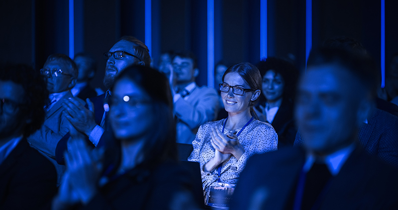 Audience members, dressed in business attire, sit and applaud in a dimly lit auditorium with blue lighting. A woman in the center, wearing glasses and a light shirt, is illuminated and clapping. Will AI replace event planners in future?