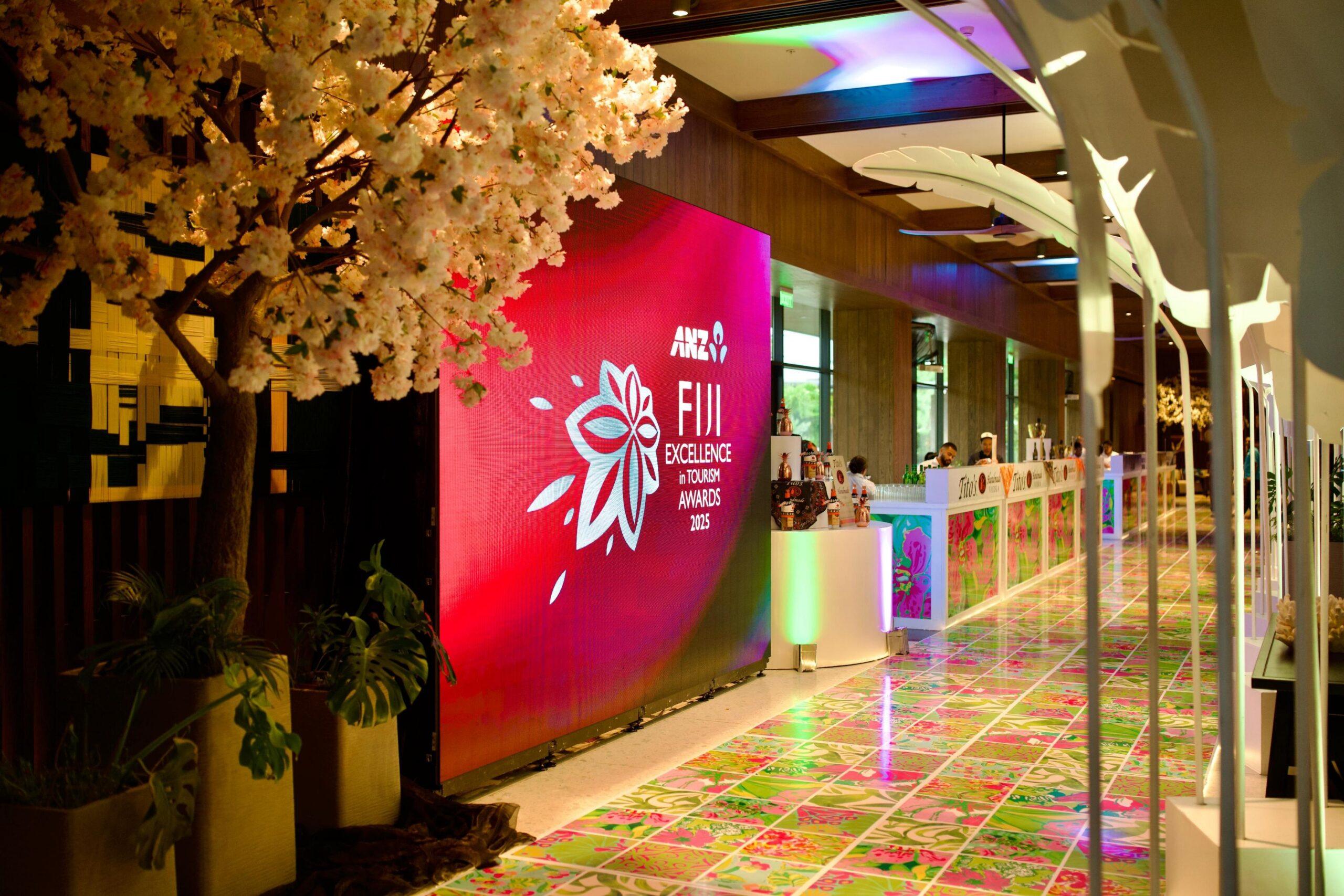 A decorated event space with a large digital screen displaying Fiji Excellence in Tourism Awards 2023, where Encore was recognised with the Bronze Award, pink flowering tree, and a colorful tiled floor with floral patterns under bright lighting.