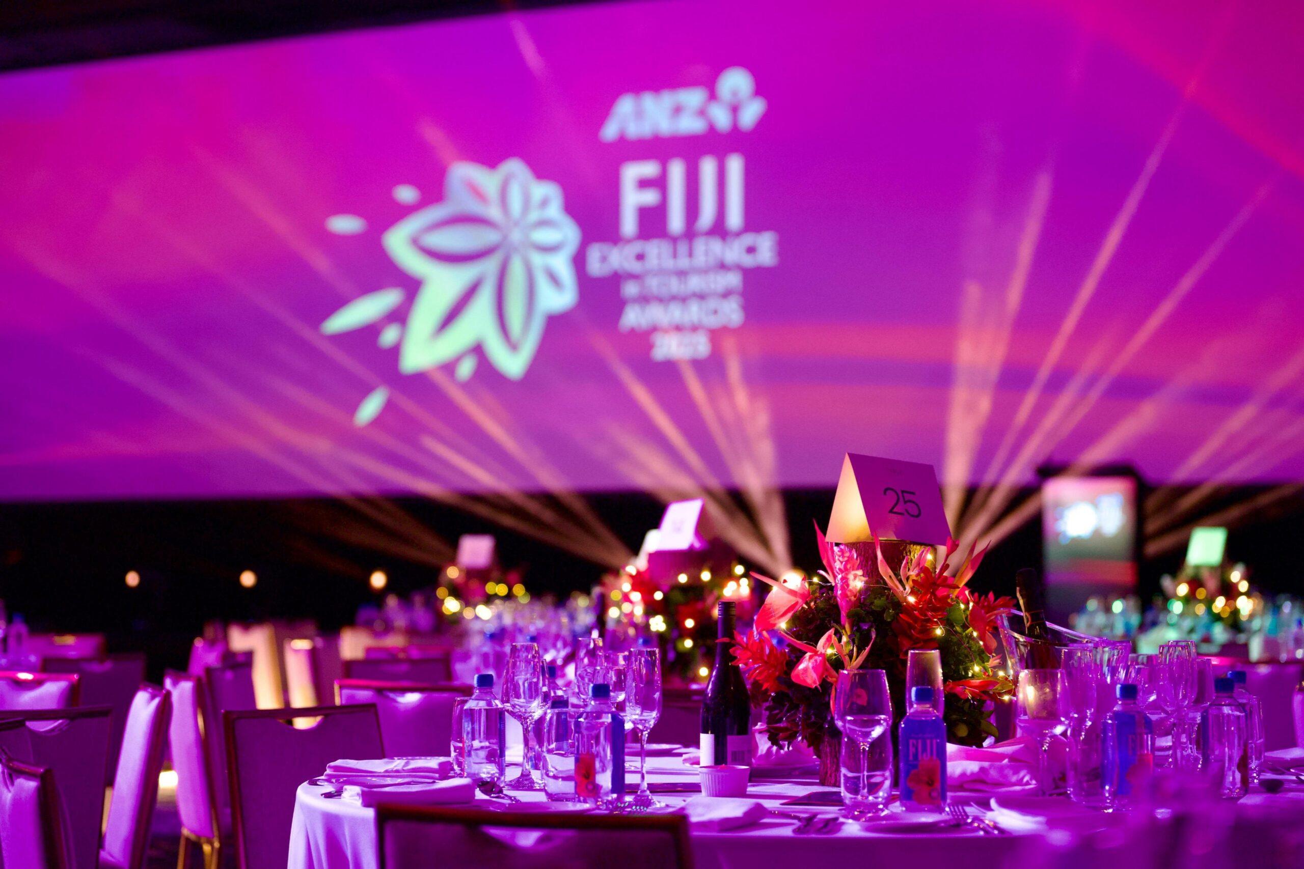 A banquet hall with elegantly set tables, floral centerpieces, and purple lighting. The backdrop displays Fiji Excellence in Tourism Awards 2015 as Encore is recognised with a Bronze Award, alongside a flower logo and ANZ branding.