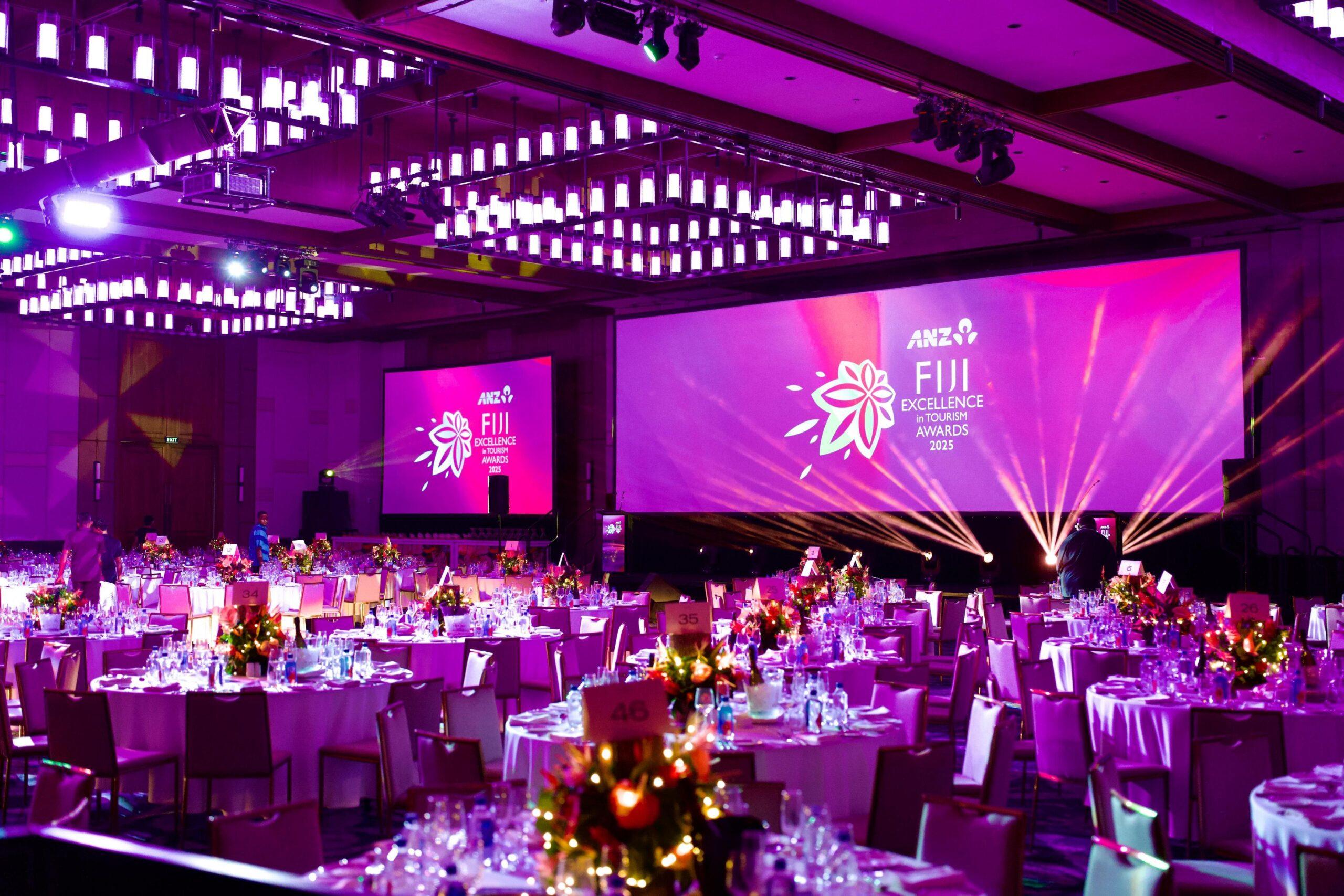 A large banquet hall decorated with round tables set for a formal event, featuring bright purple lighting and a large screen displaying ANZ Fiji Excellence Awards 2023, where Encore was recognised with a Bronze Award at the Fiji Excellence in Tourism Awards.