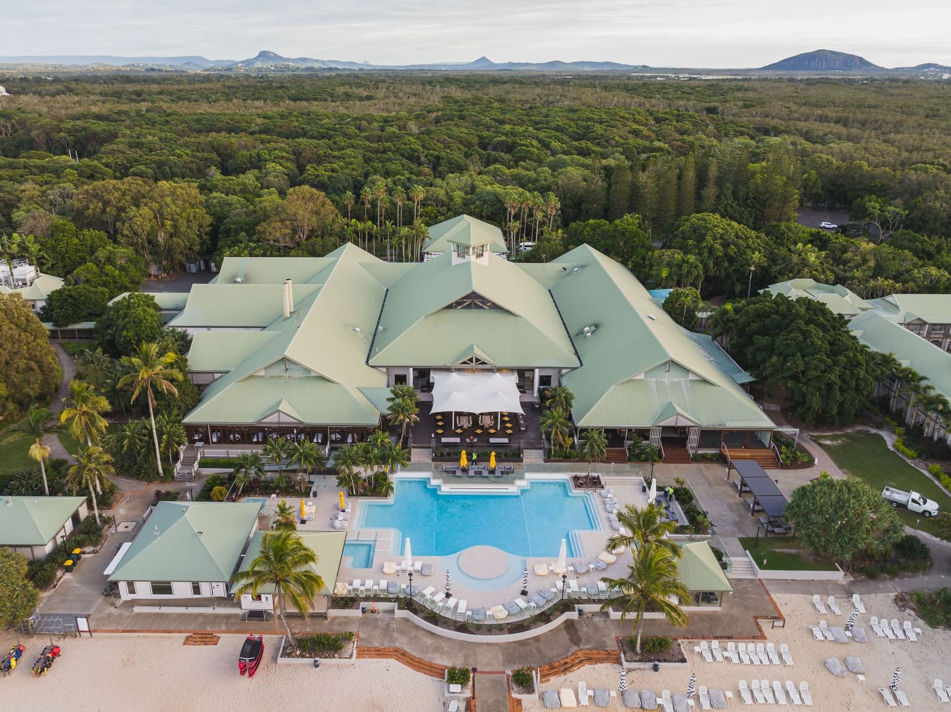 A large resort with green roofs, a central pool, sun loungers, and palm trees is surrounded by dense forest and sandy beach, viewed from above. Mountains are visible in the distance—this is Novotel Sunshine Coast at its finest.