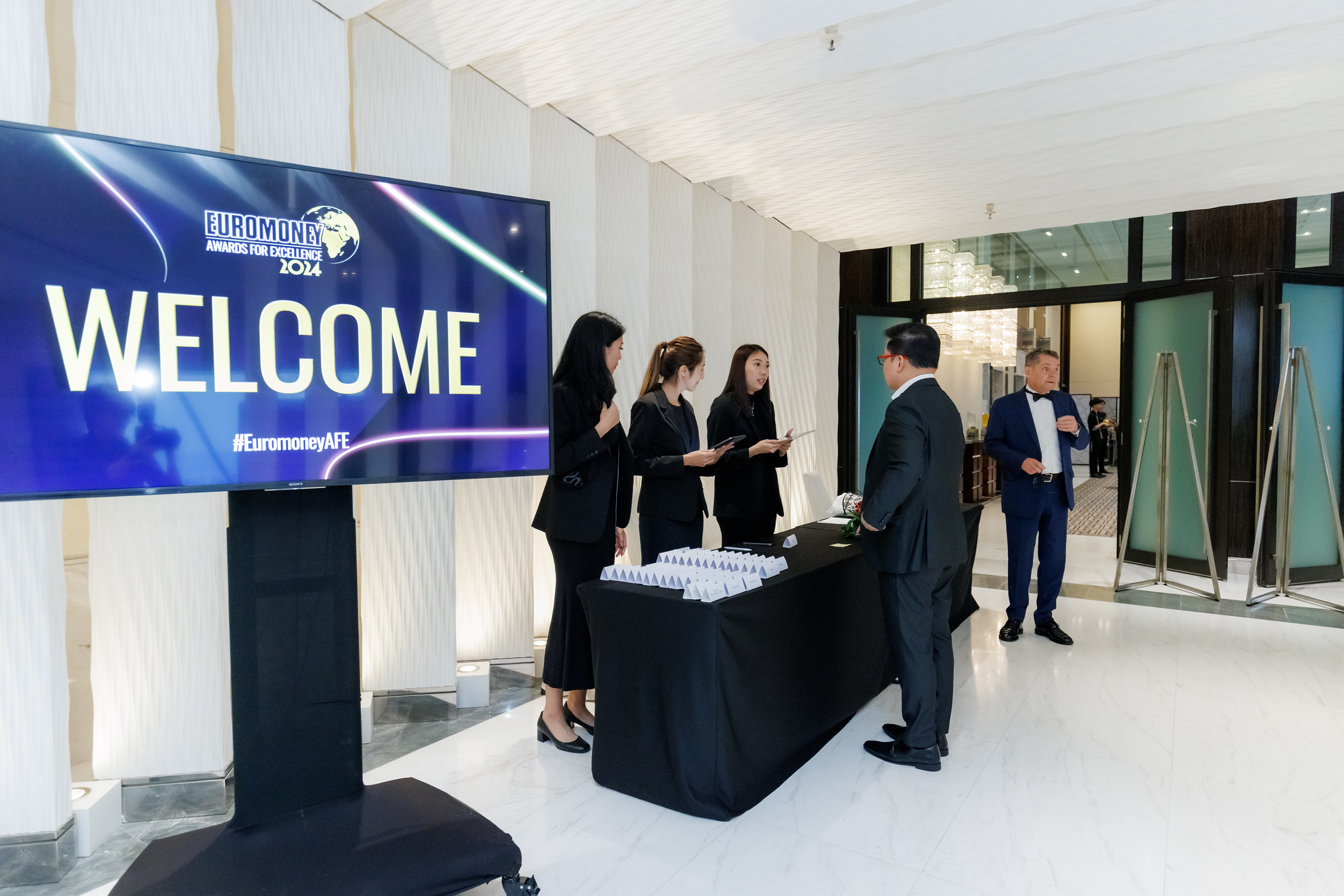 Four women at a check-in table greet two men in suits at a formal event; a large screen beside them displays “WELCOME” and “Euromoney Asia-Pacific Awards for Excellence 2024” in a modern, white, well-lit venue.