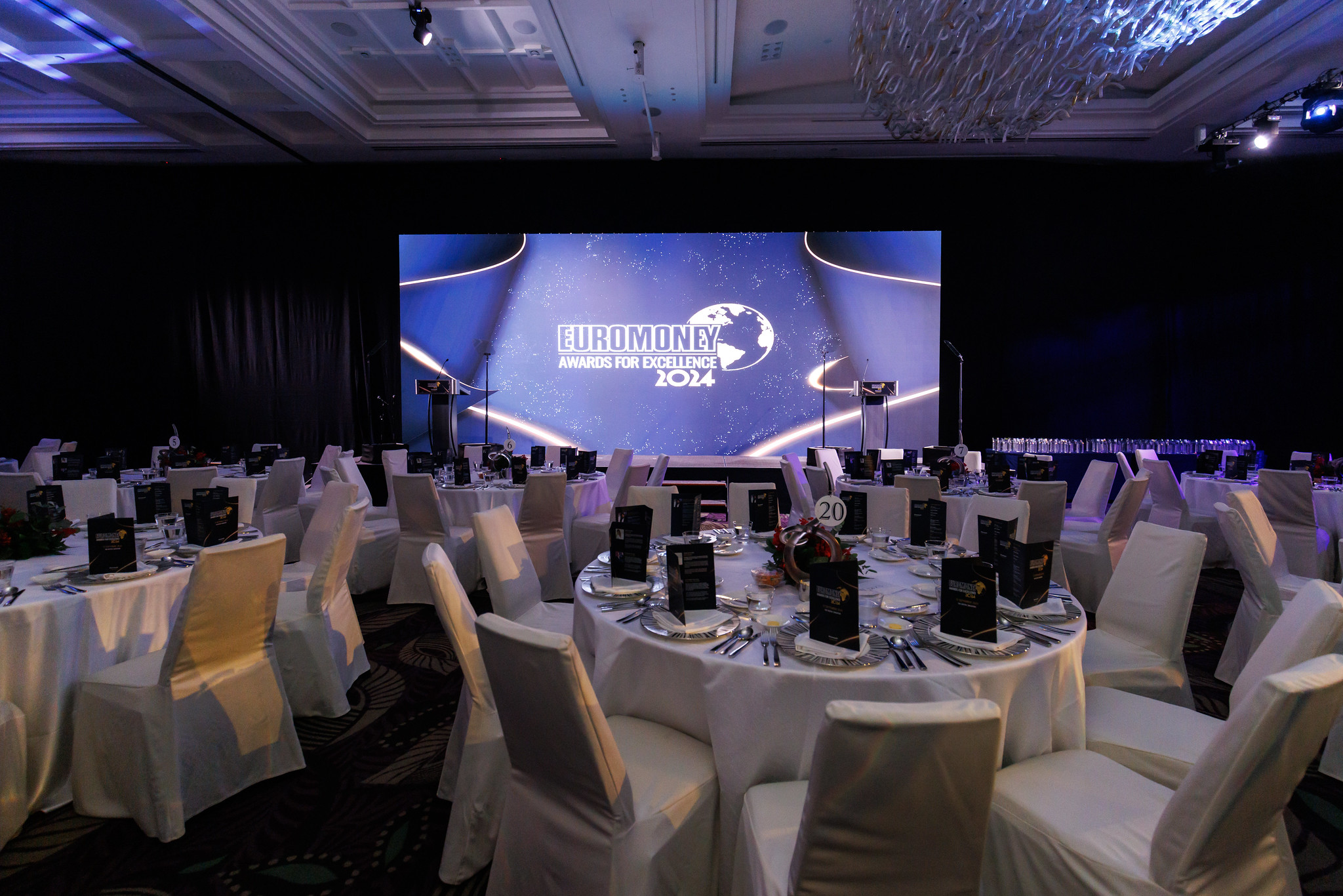 A formal event setup with round tables draped in white cloths and chairs, facing a large screen displaying Euromoney Asia-Pacific Awards 2024 in a dimly lit banquet hall. (Euromoney Asia-Pacific Awards for Excellence 2024 - The Westin Singapore x Encore)