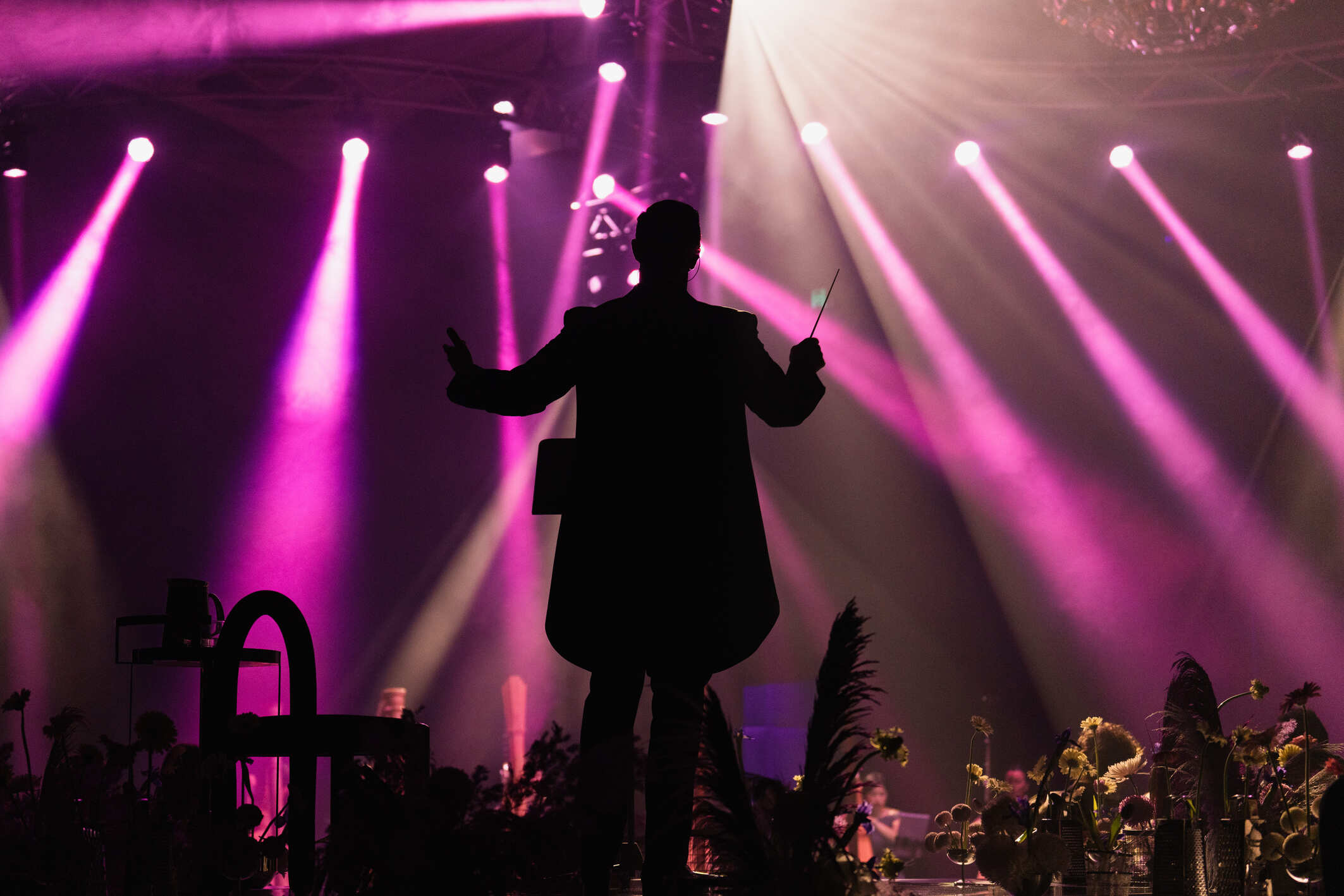 A conductor stands on stage with arms raised, silhouetted against dramatic purple and pink stage lights, leading an orchestra during a live performance. X as the Unknown Variable Showcase at Crown Melbourne - an Encore Creative Production. Reimagining the palladium during AIME