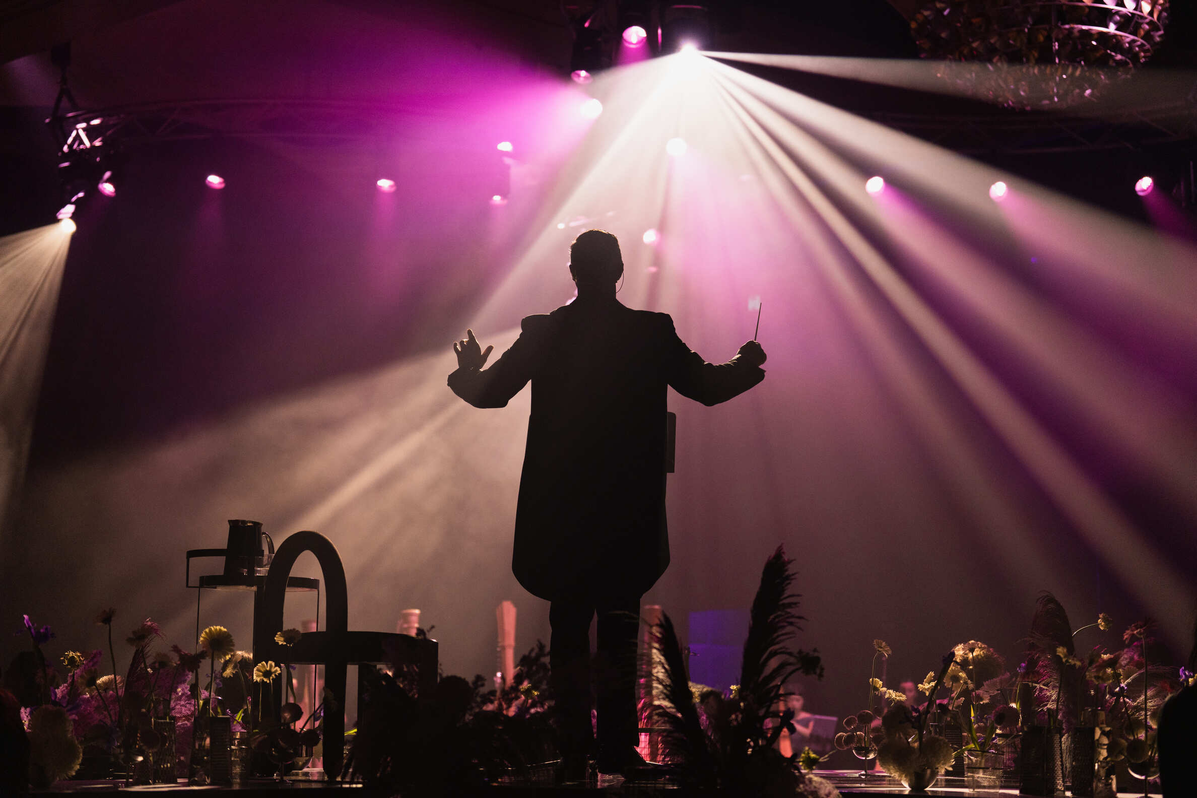 A conductor stands on stage with arms raised, silhouetted against dramatic purple and white spotlights, while musicians sit and flowers decorate the foreground.