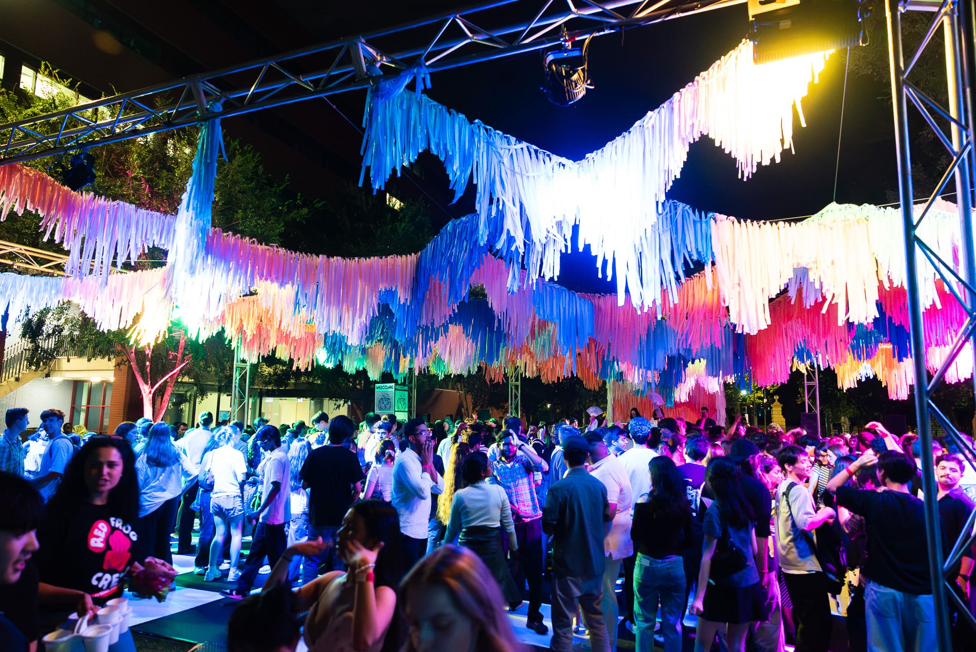 A lively crowd gathers under colorful, hanging streamers illuminated by vibrant lights at the AUSA Welcome Party, an outdoor night event surrounded by trees at Adelaide University.