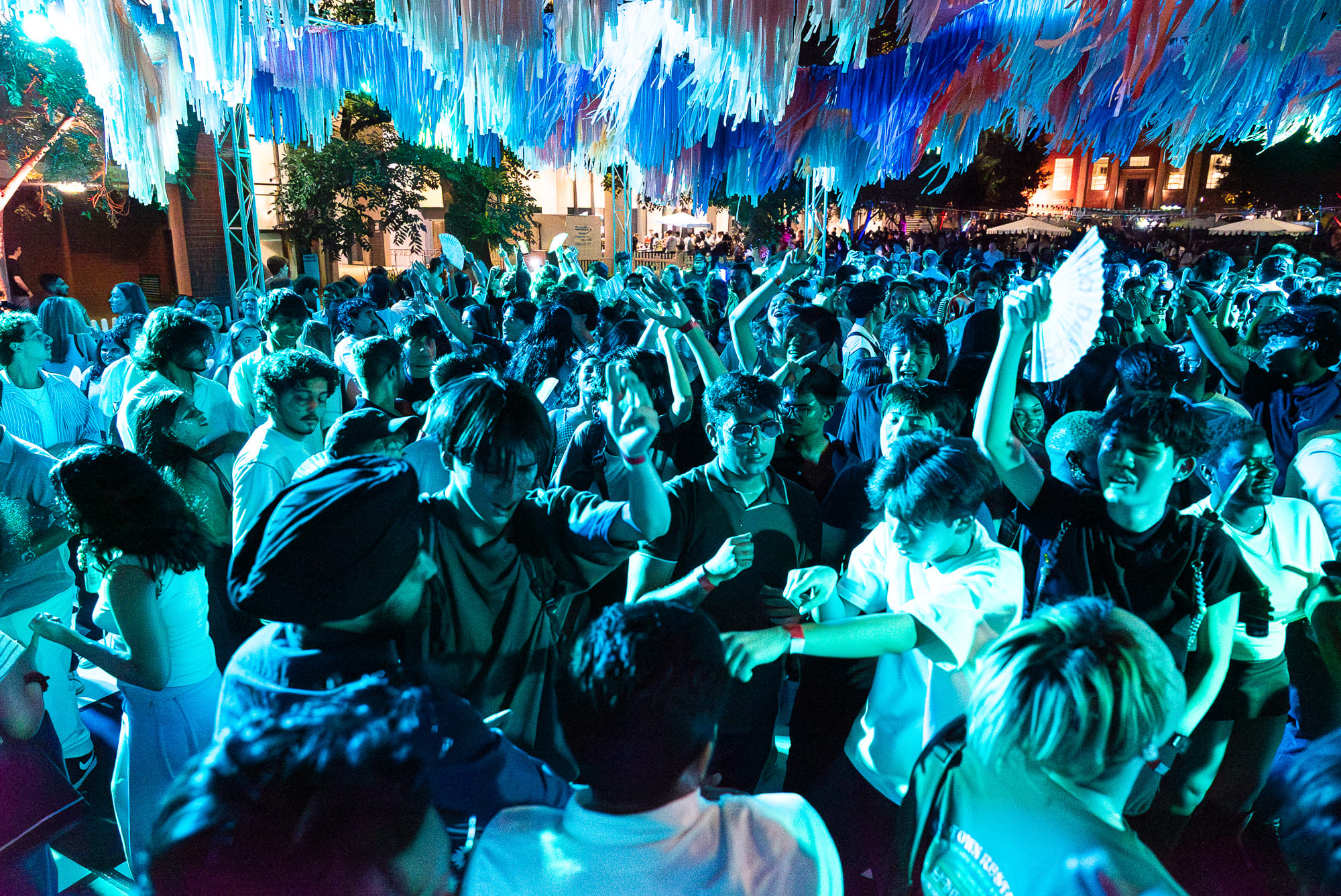 A large crowd of young people dances energetically under colorful, hanging decorations at a vibrant outdoor night party, illuminated by blue and purple lights. AUSA Adelaide Welcome Party - (8)