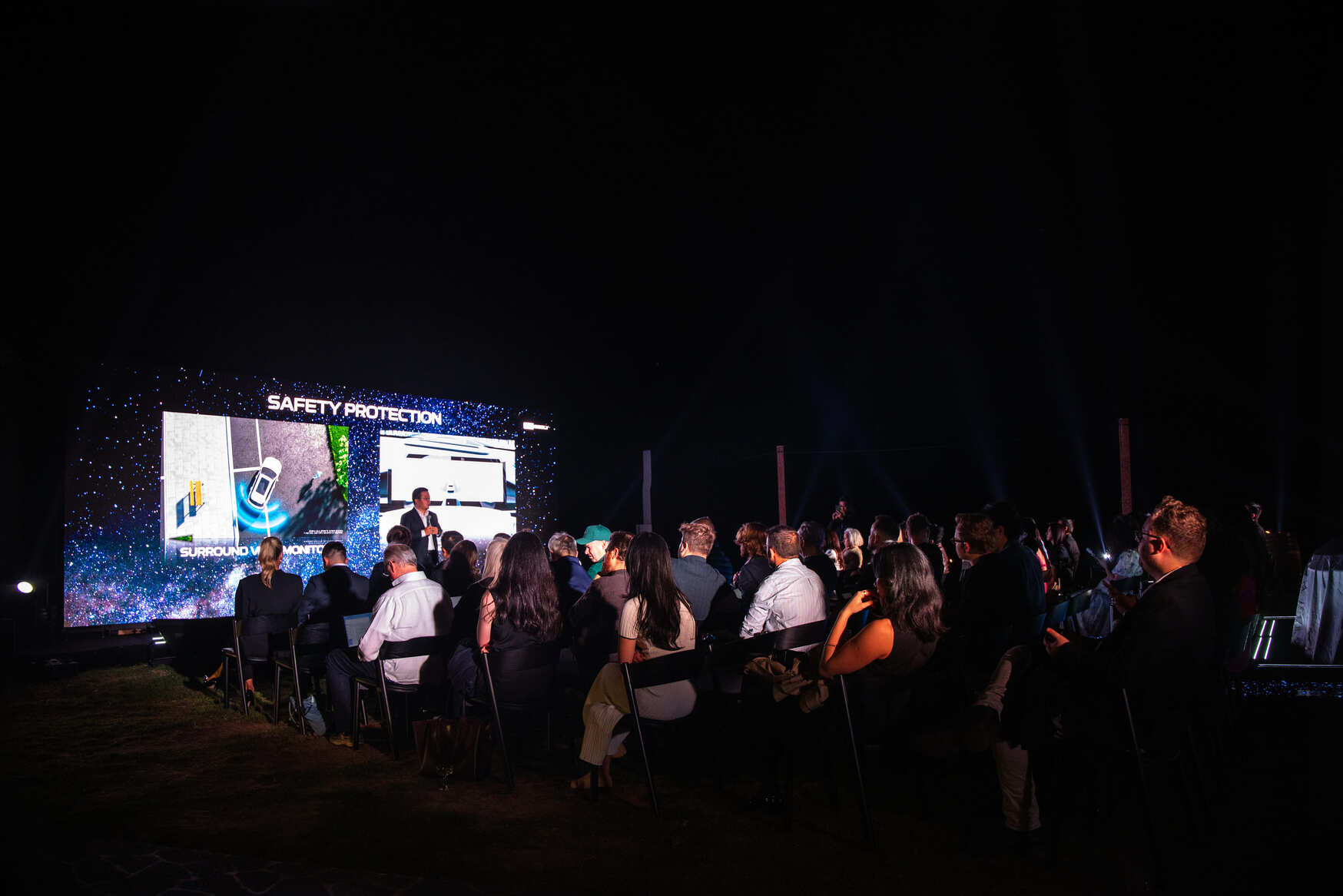 A group of people sits outdoors at night, watching a large screen displaying a presentation titled SAFETY PROTECTION. The audience is dressed formally and seated in rows of chairs.