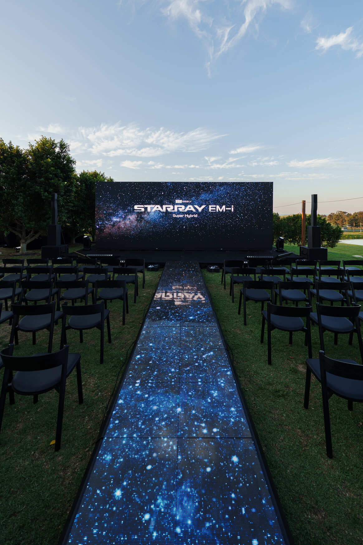 An outdoor event setup with rows of black chairs facing a stage and a large screen displaying STARRAY EM-H against a starry background. The walkway leading to the stage is illuminated with a matching starry galaxy design.