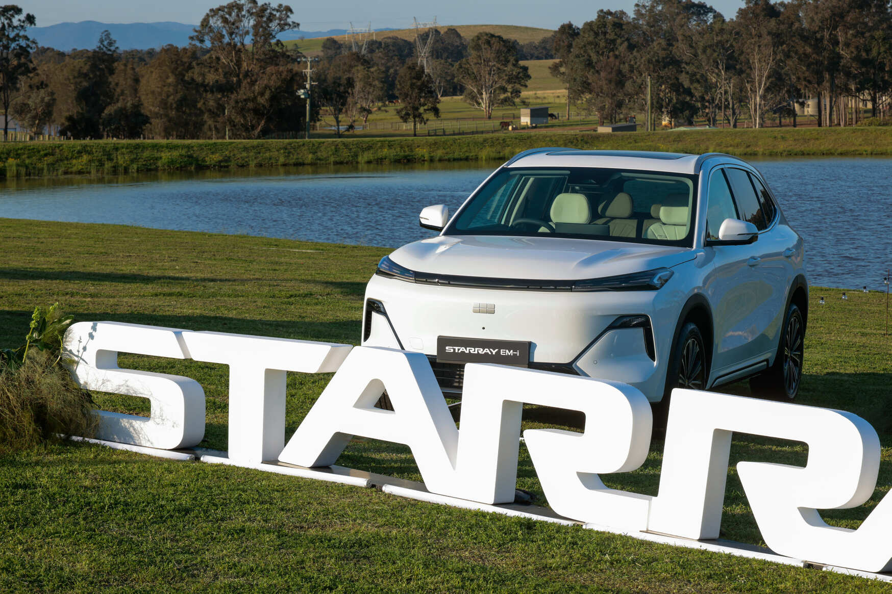 A white SUV labeled STARRAY 6M4 is parked on grass beside a lake, with large white 3D letters spelling STARR in front. Trees, hills, and water are visible in the background under a clear sky.