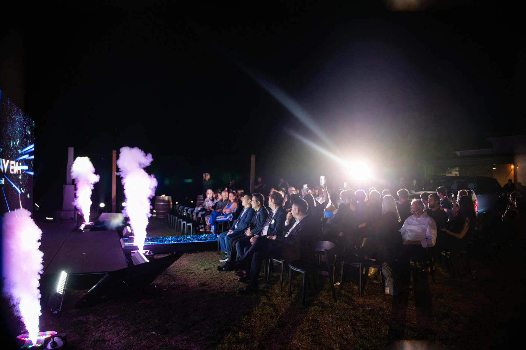 An audience sits outdoors at night, watching a brightly lit stage with smoke effects and colorful lighting. People are seated in rows, some illuminated by stage lights, creating a dramatic atmosphere.