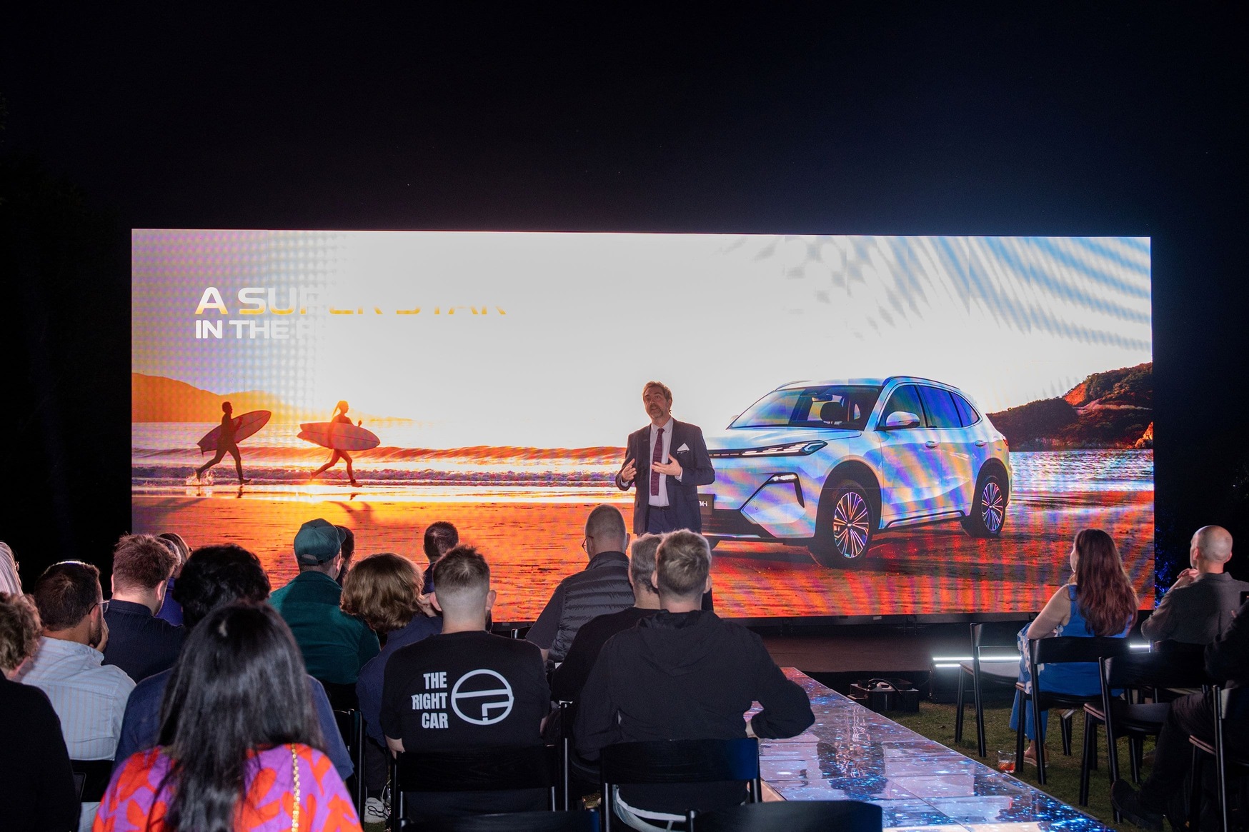 A speaker presents in front of an audience, with a large screen behind showing a car on a beach and two surfers walking, under the partially visible text A SUN... IN THE....
