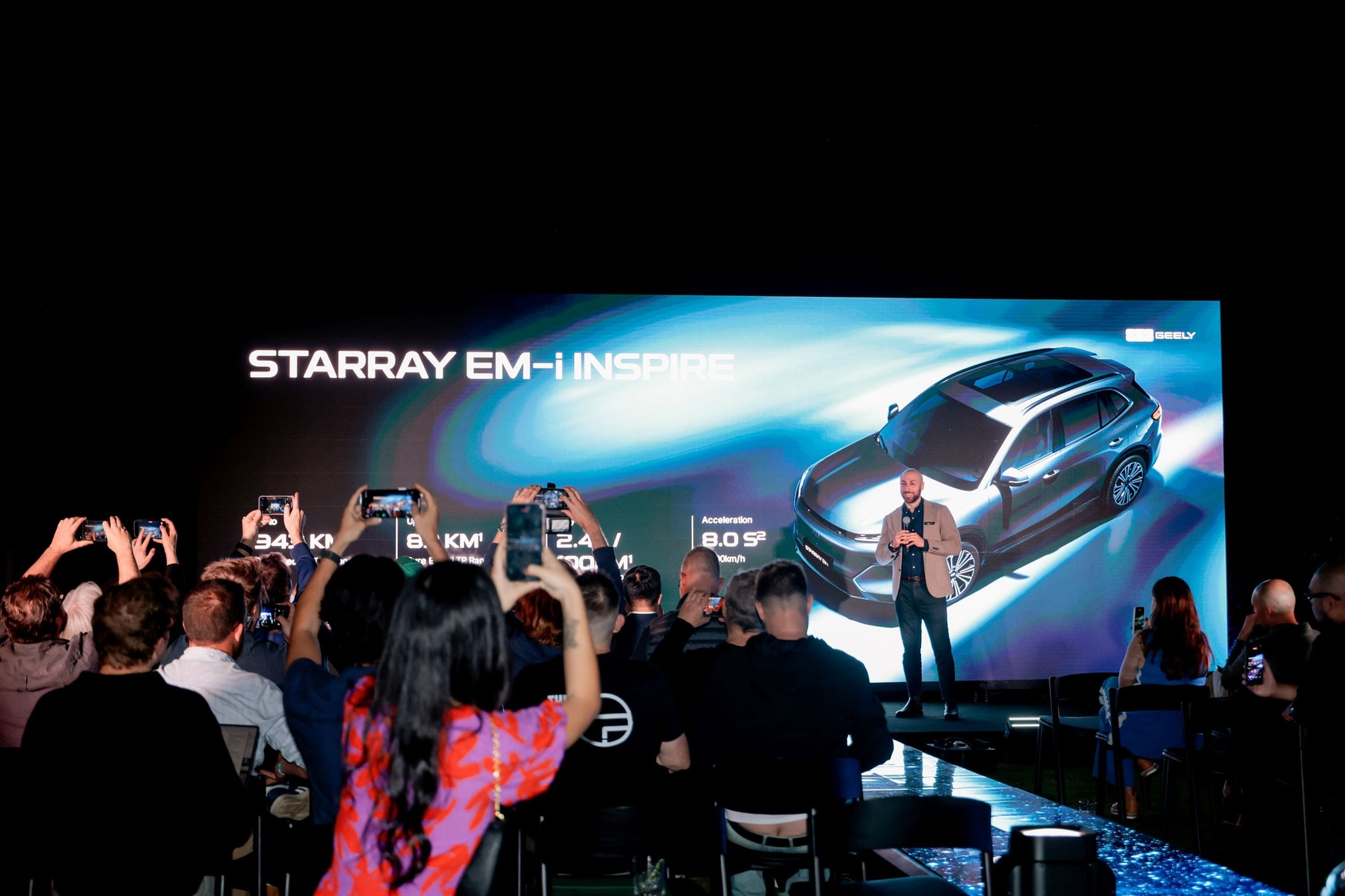 A man stands on stage presenting the STARRAY EM-i INSPIRE car to an audience, with people taking photos and a large screen displaying the car’s image and features in the background.