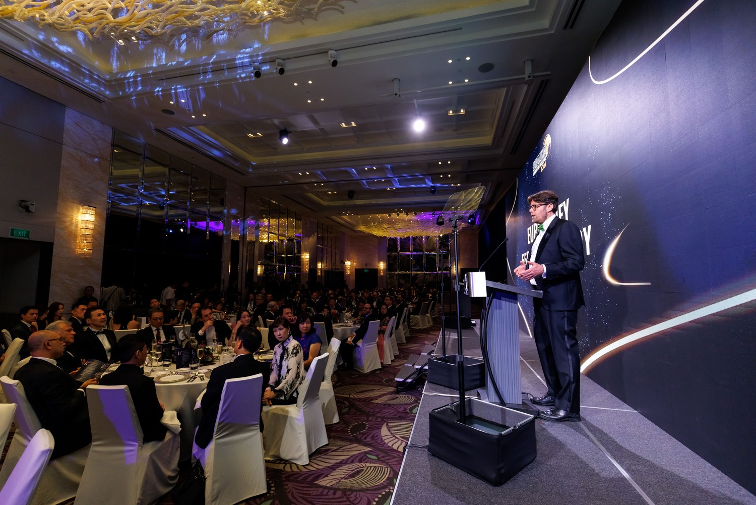 A man in a suit speaks at a podium on stage during a formal event in a large, elegant banquet hall filled with seated guests at round tables. The room is brightly lit with decorative ceiling lights.