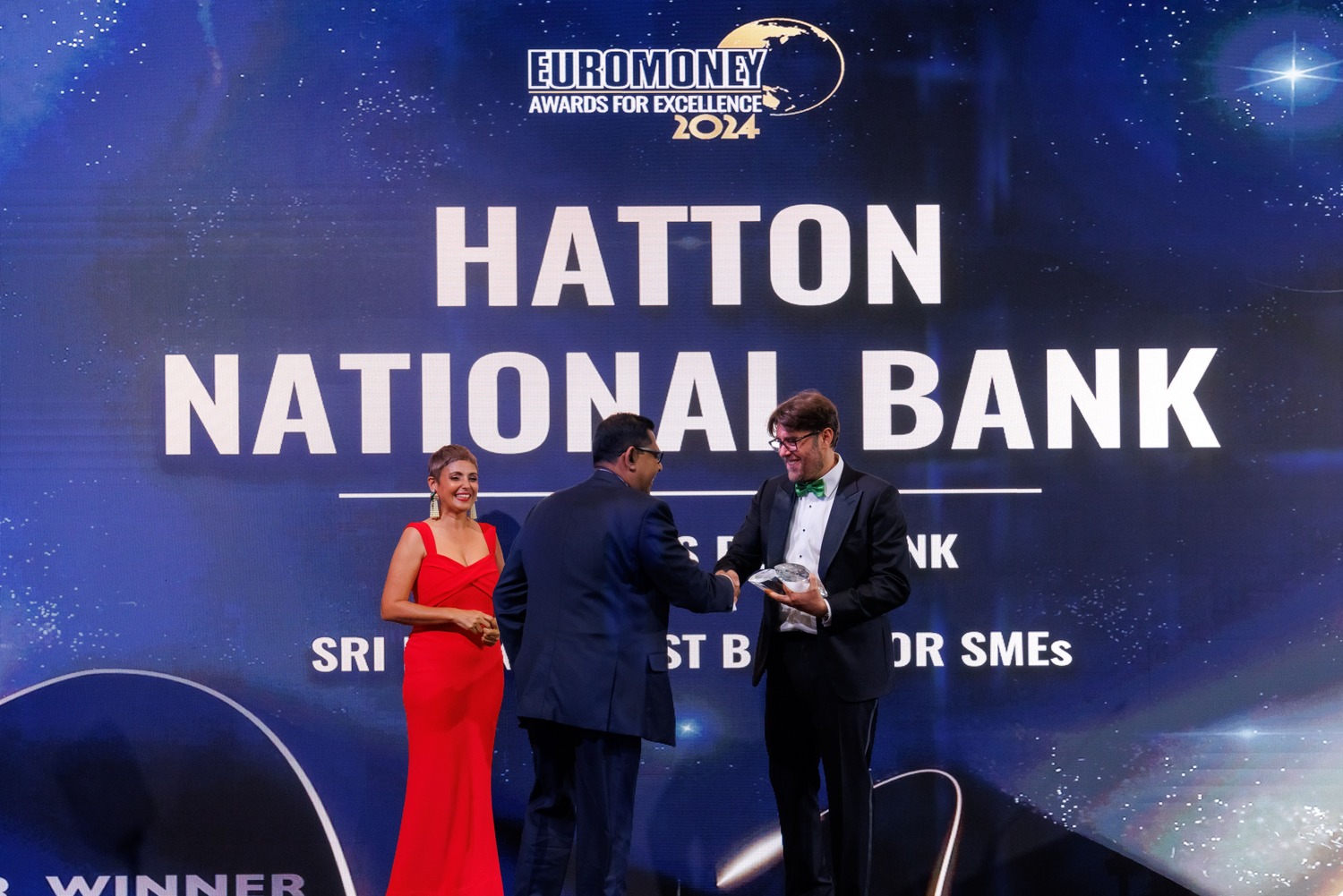 Three people stand on stage at the Euromoney Awards for Excellence 2024; a woman in a red dress smiles as two men in suits shake hands in front of a backdrop reading Hatton National Bank: Sri Lanka’s Best Bank for SMEs.