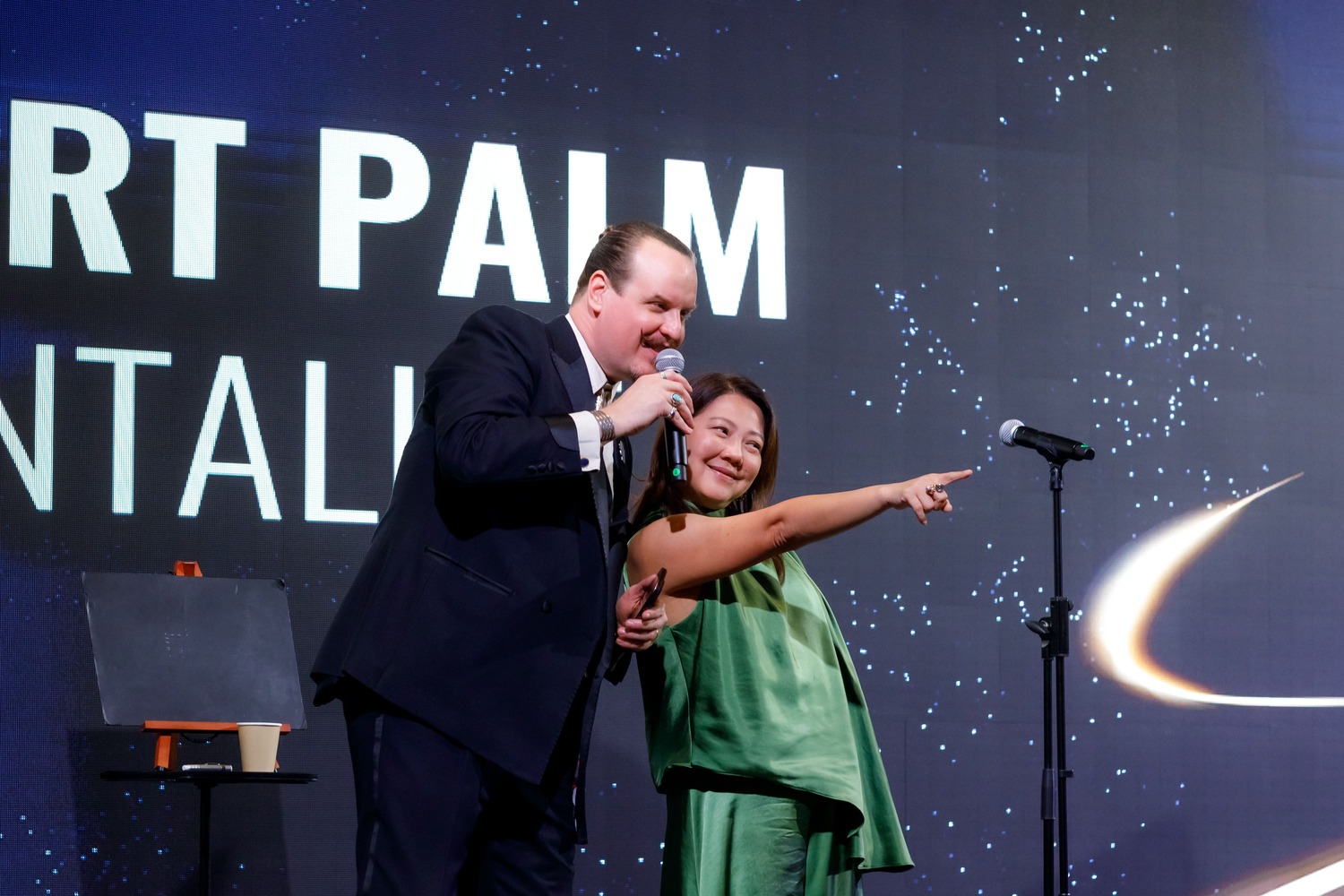 A man in a suit holds a microphone and smiles next to a woman in a green dress who is pointing excitedly, both standing on stage in front of a large screen displaying text and a starry background.