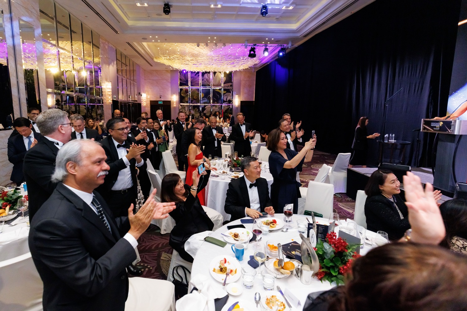 A crowd of people in formal attire stand and clap around decorated round tables at a banquet event in a large, elegant hall with high ceilings and bright lighting.