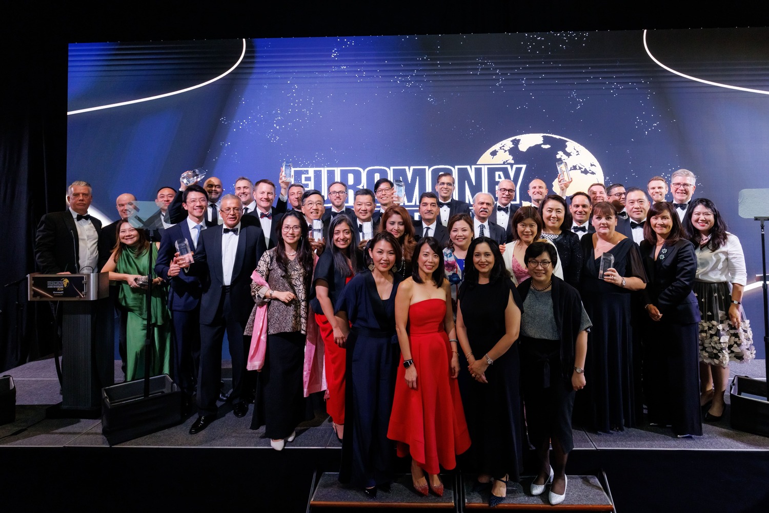 A large group of people dressed in formal attire poses and smiles on stage at an awards event, with a backdrop displaying the word EUROMONEY and an image of the globe. Euromoney Asia-Pacific Awards for Excellence 2024 - The Westin Singapore x Encore