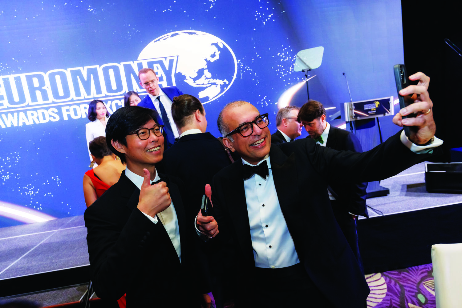 Two men in formal wear take a selfie and smile at an awards event. A large screen behind them displays Euromoney Awards for Excellence with a globe graphic. Other attendees are visible in the background. Euromoney Asia-Pacific Awards for Excellence 2024 - The Westin Singapore x Encore