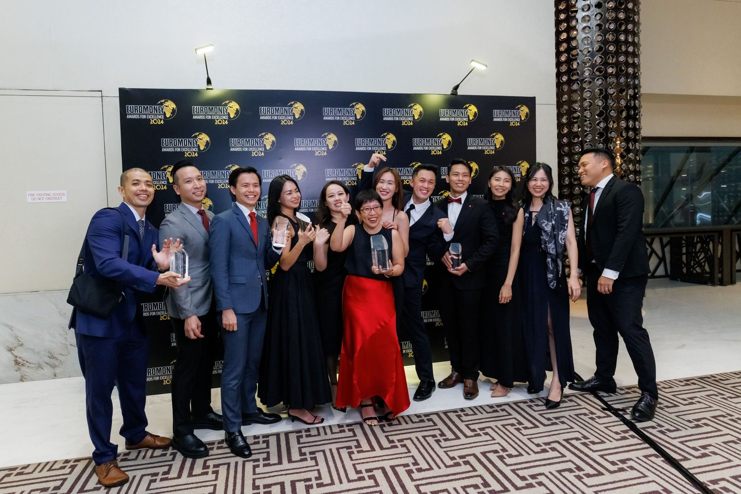 A group of twelve people in formal attire pose together, smiling and holding glass awards in front of a black step-and-repeat backdrop at an indoor event.