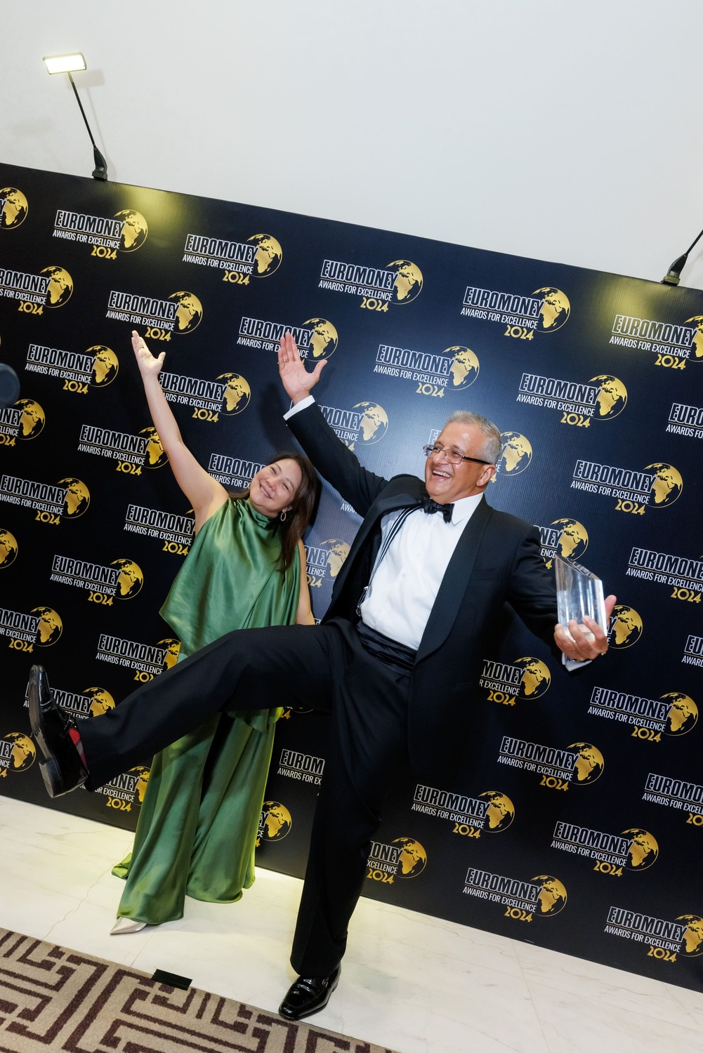 A woman in a green dress and a man in a tuxedo holding an award pose playfully with wide smiles and raised arms and legs in front of a backdrop with Euromoney 2024 logos.