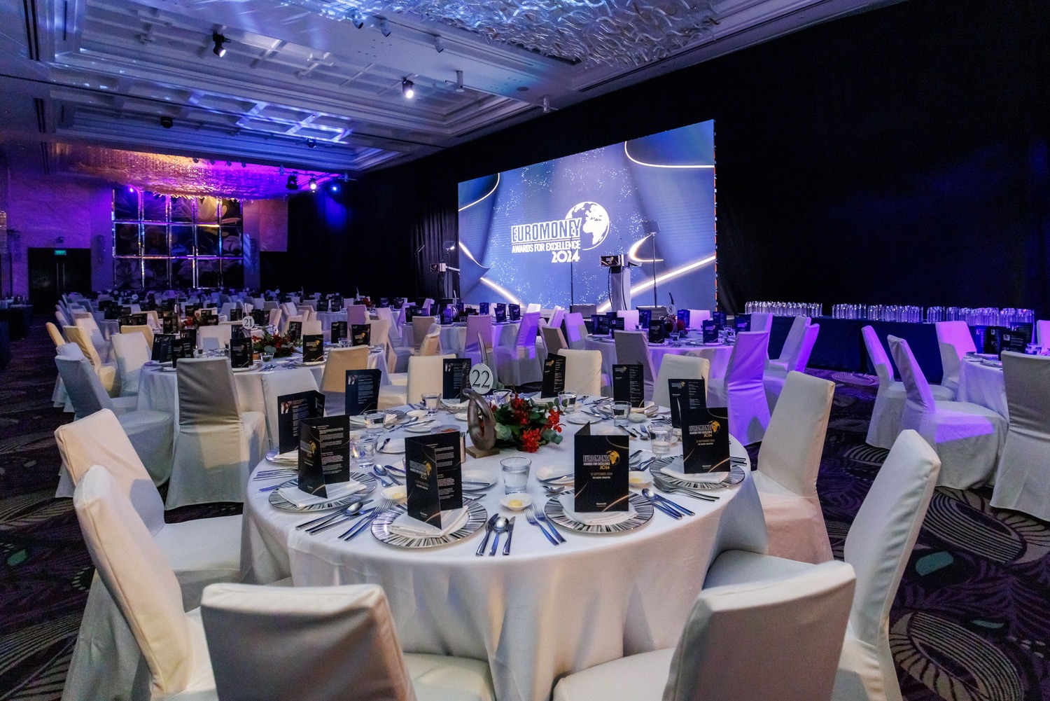 A large, elegant banquet hall set up for an event, with round tables covered in white tablecloths, surrounded by white chairs, and a large screen at the front displaying HARMONY AWARDS 2024.