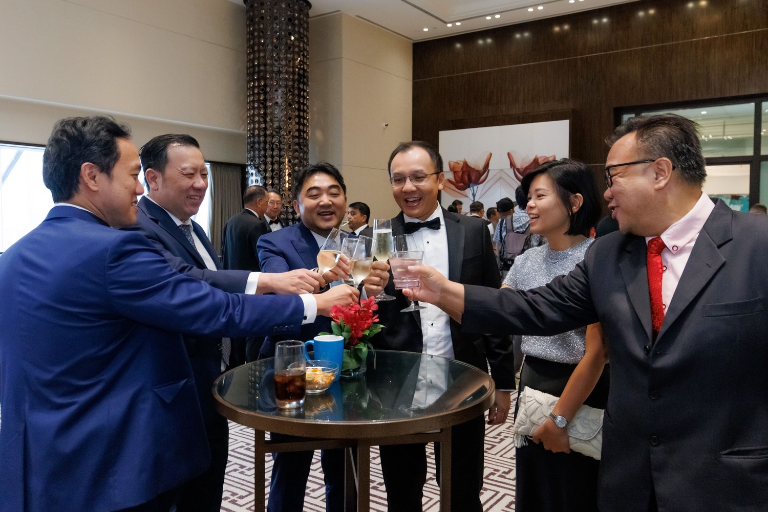Six people dressed in formal attire are standing around a tall table, smiling and raising their glasses in a toast at a lively indoor event. The background shows other guests and elegant decor.