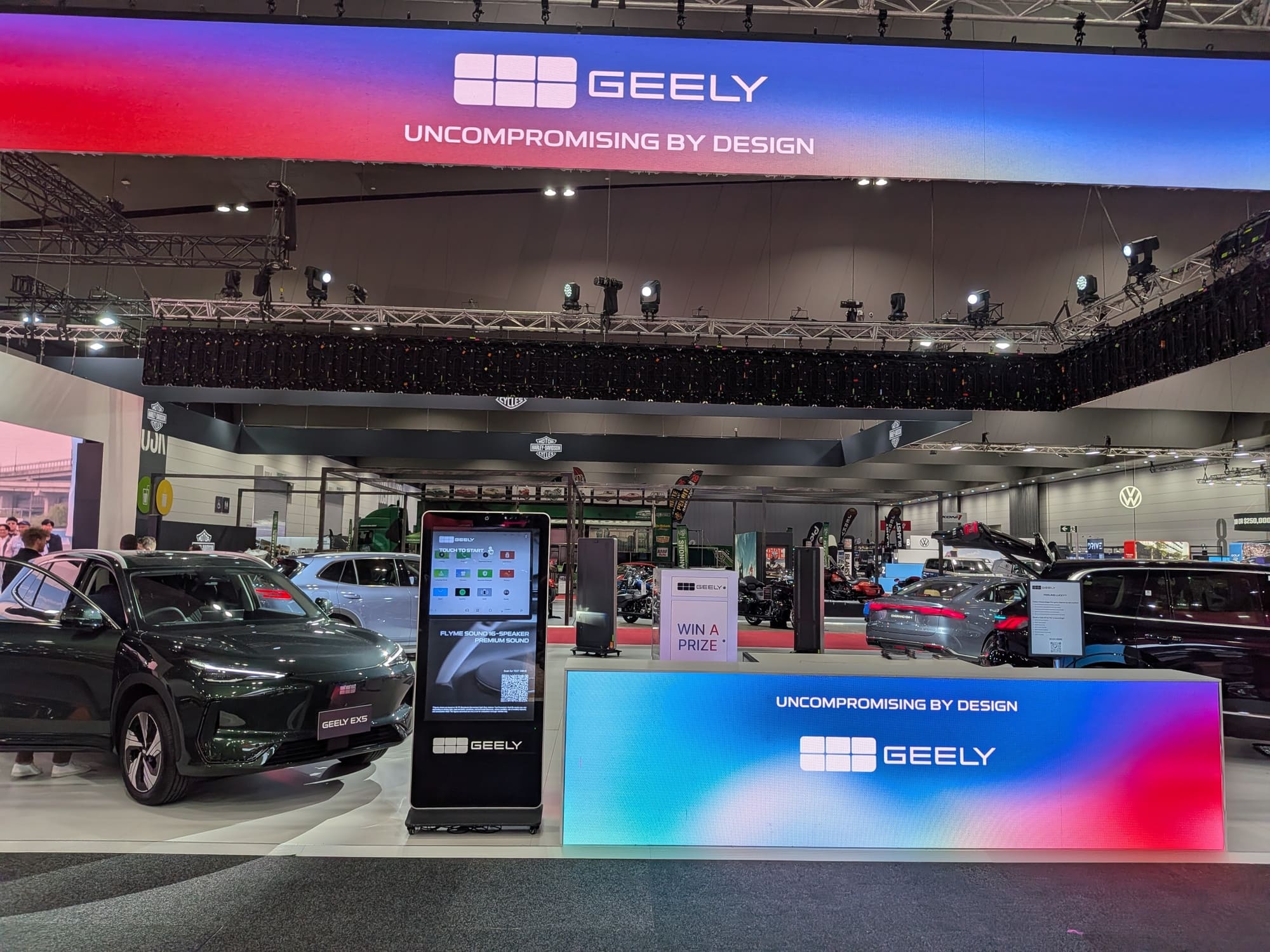 Geely stand featuring digital display kiosk, illuminated counter and vehicles in background - Encore Event Technologies