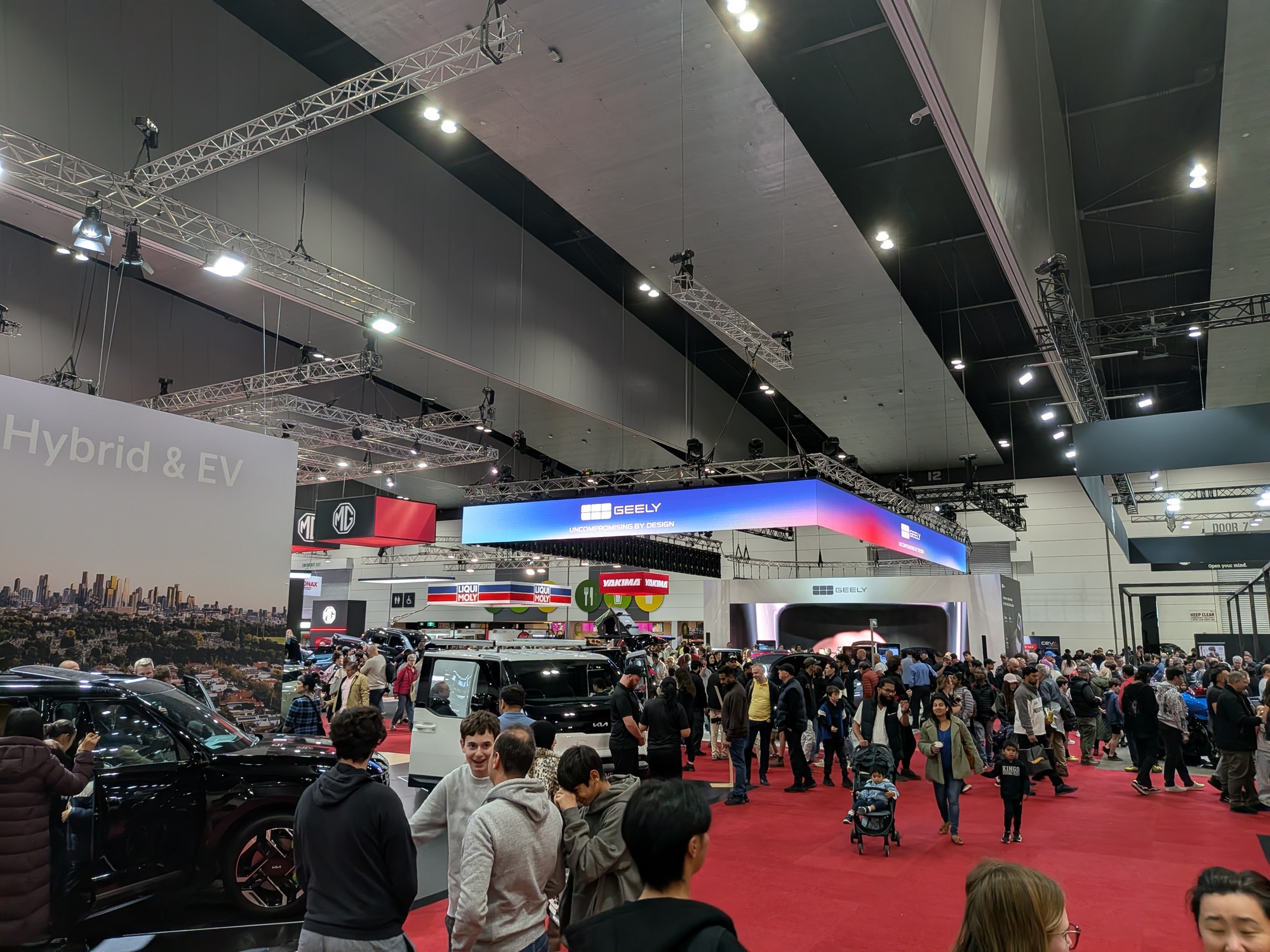 Wide view of busy motor show floor with Geely exhibition stand, overhead lighting rig and large crowds on red carpet - Encore Event Technologies