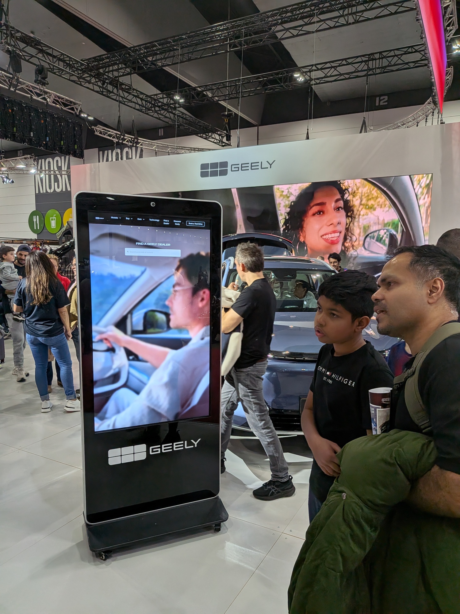 Interactive Geely touchscreen kiosk in foreground with attendees viewing digital content and vehicles behind - Encore Event Technologies
