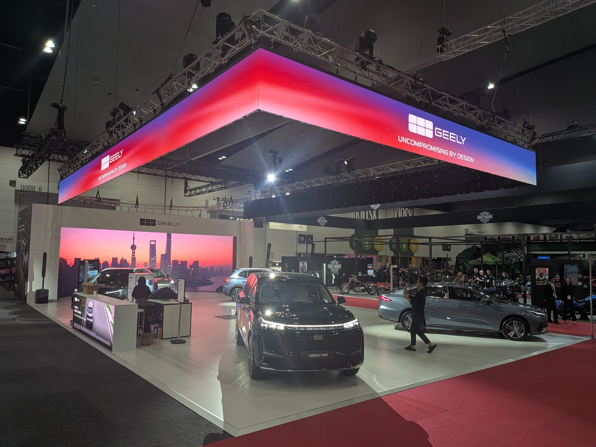 Geely exhibition stand with overhead illuminated branding cube, LED video wall backdrop and multiple vehicles showcased on display platform - Encore Event Technologies