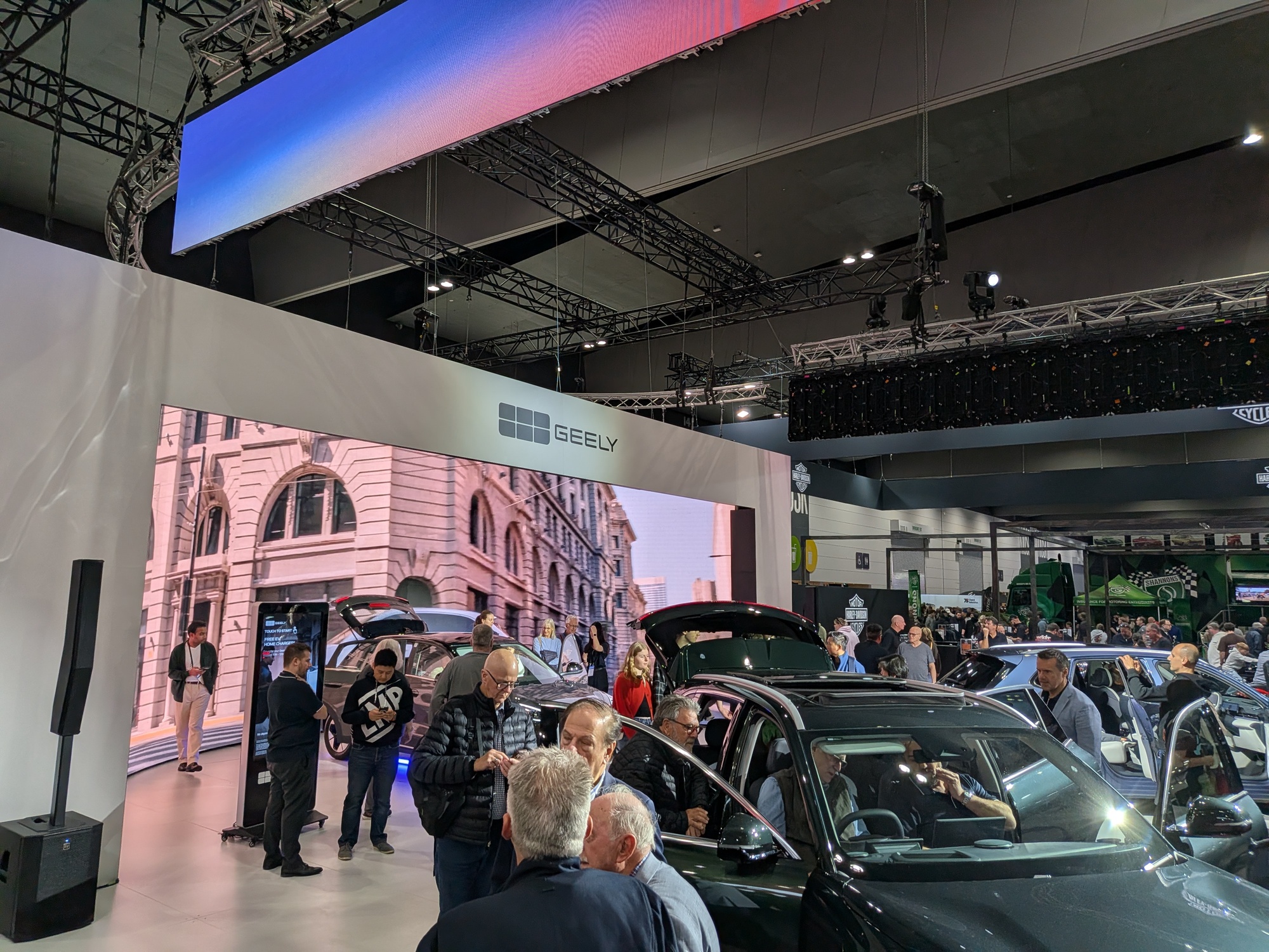 Geely exhibition stand with overhead illuminated branding cube, LED video wall backdrop and multiple vehicles showcased on display platform - Encore Event Technologies