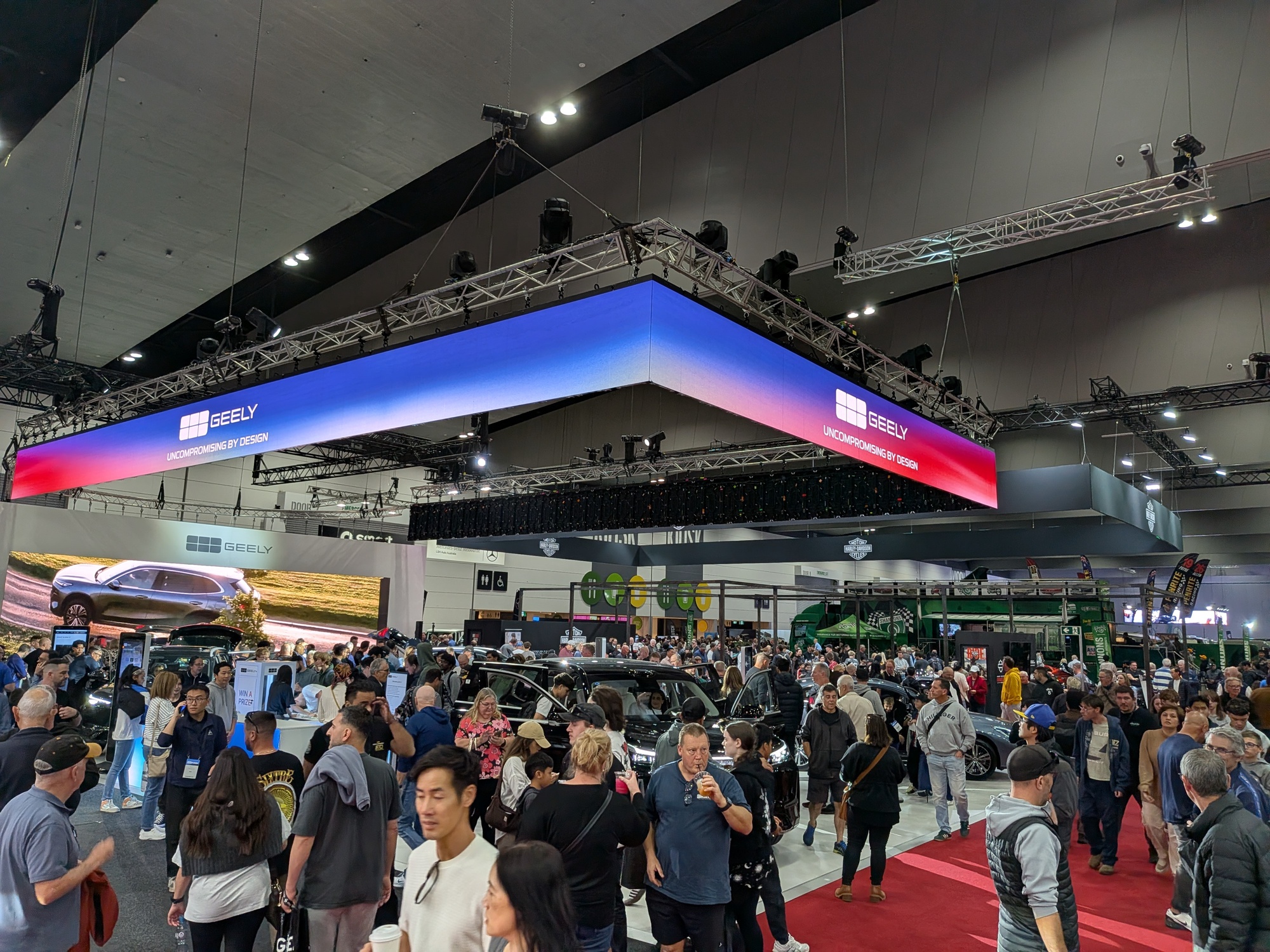 Wide exhibition floor view centred on Geely booth with illuminated overhead cube, vehicles and high visitor foot traffic - Encore Event Technologies