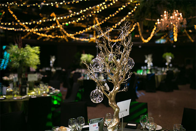 Gold tree centrepiece with hanging glass globes surrounded by banquet tables under ceiling fairy lights in a dimly lit venue – Encore Event Technologies
