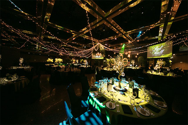 Gala dinner setup with illuminated tables, overhead string lights and dual presentation screens in a darkened event ballroom – Encore Event Technologies