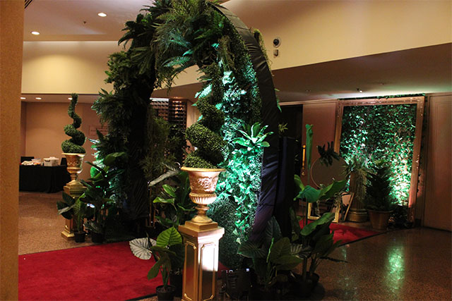 Greenery-covered event entrance arch with potted plants, uplighting and a red carpet leading into a venue space – Encore Event Technologies