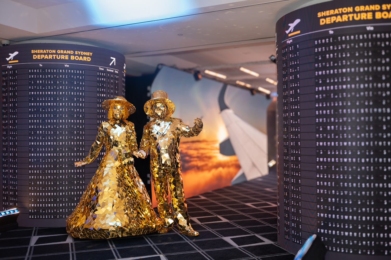 Gold mirrored performer sculptures positioned between illuminated airport-style departure boards within a travel-themed event space at Sheraton Grand Sydney - Encore Event Technologies
